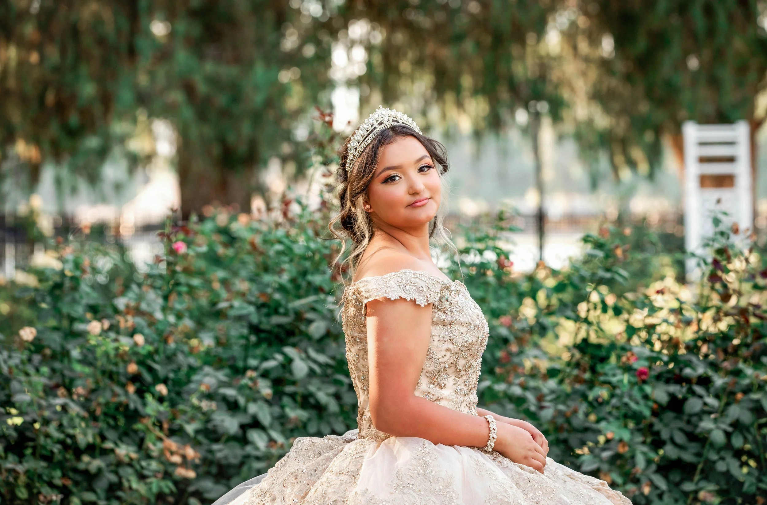 A young woman in an ornate, off-the-shoulder, cream-colored gown with intricate beading and lace, sitting outdoors among greenery and pink flowers, wearing a tiara and jewelry, with a soft smile on her face.
