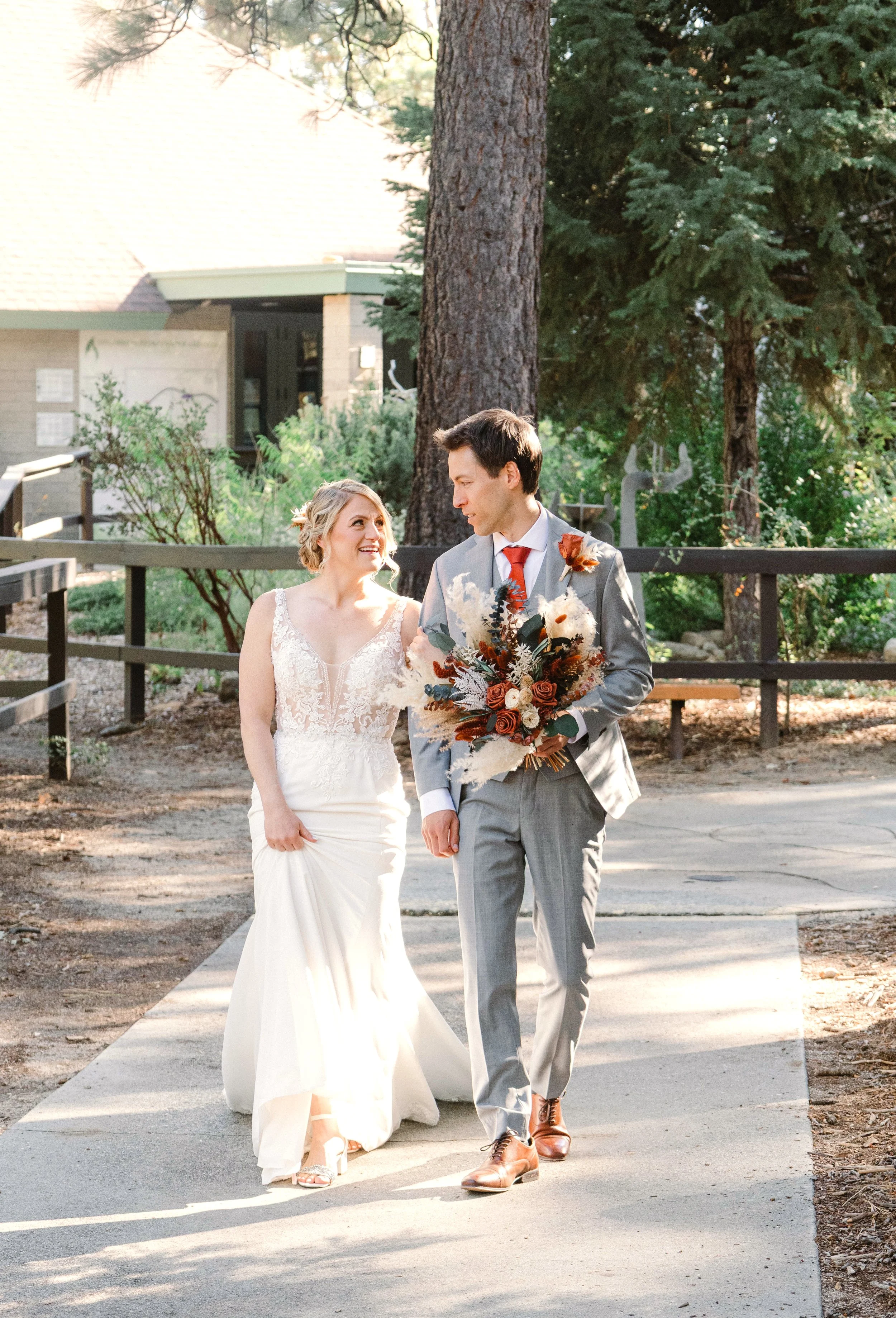 A bride and groom walking outdoors, smiling at each other. The bride is wearing a lace wedding gown and heels, and the groom is in a light gray suit with a red tie, holding a bouquet of orange and white flowers. They are surrounded by trees and greenery, with sunlight casting a warm glow.