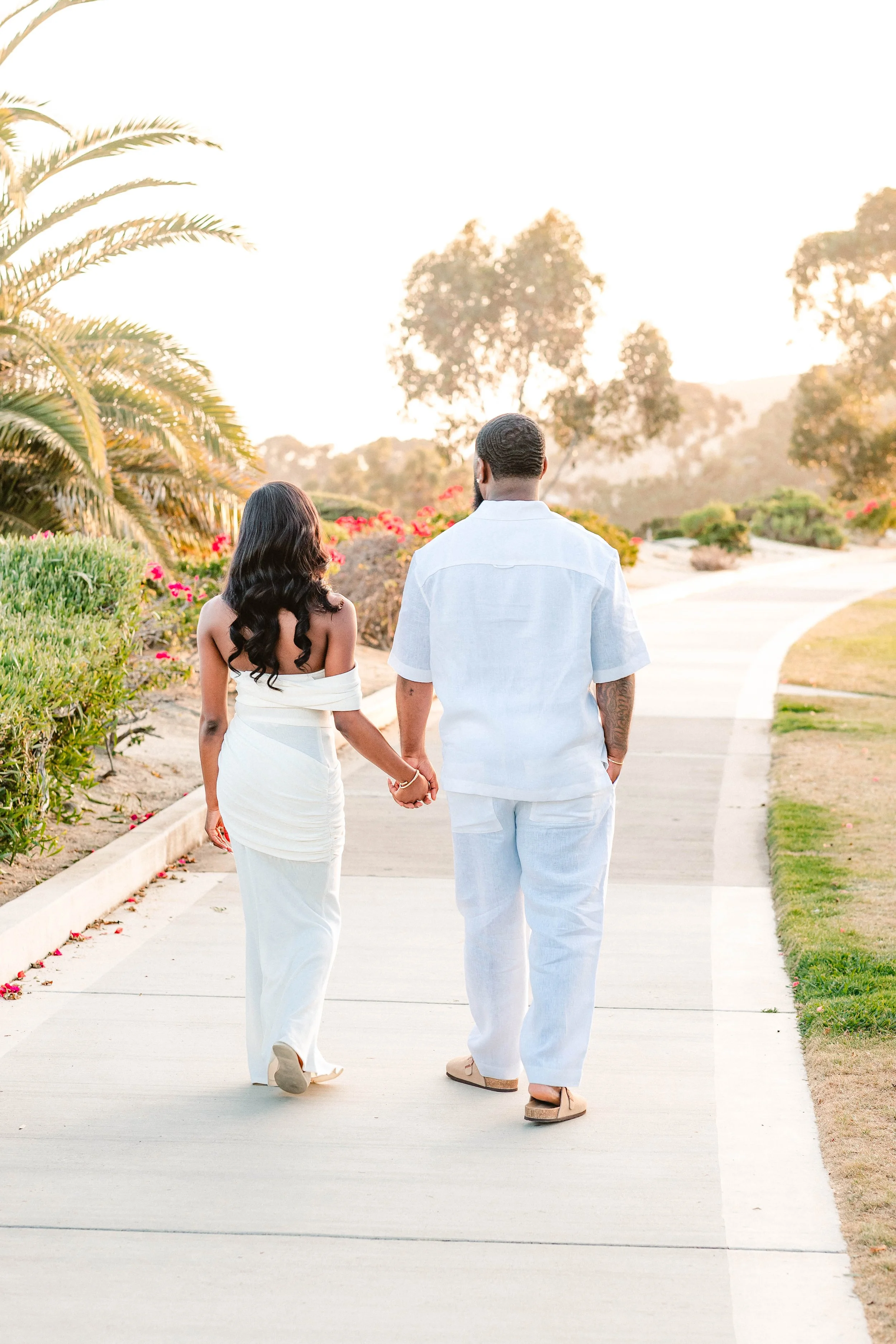 A couple walking hand-in-hand on a paved path in a garden during sunset, both dressed in white.