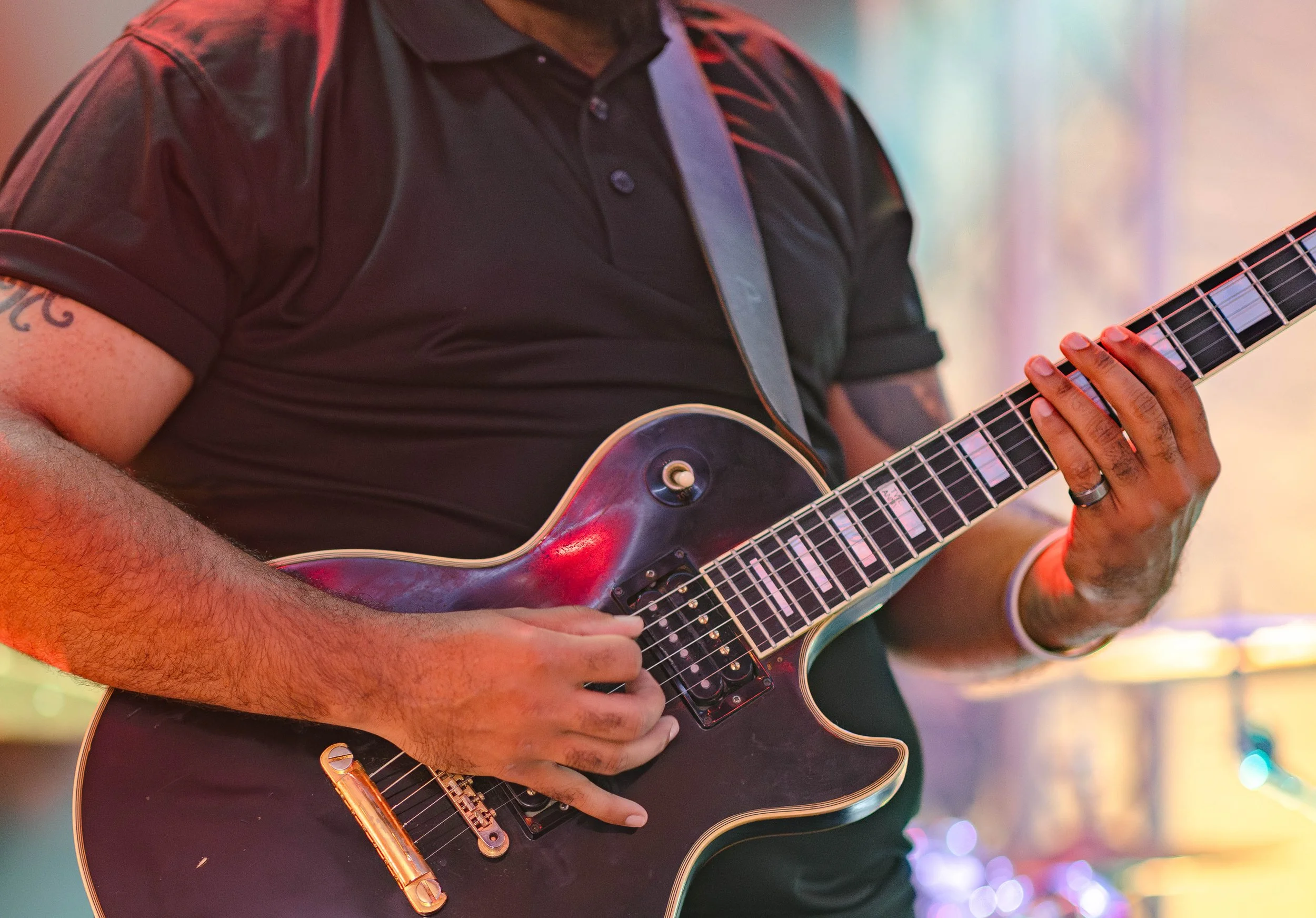 Close-up of a person playing an electric guitar, wearing a black shirt, tattooed arm, and wedding ring, with colorful lights in the background.
