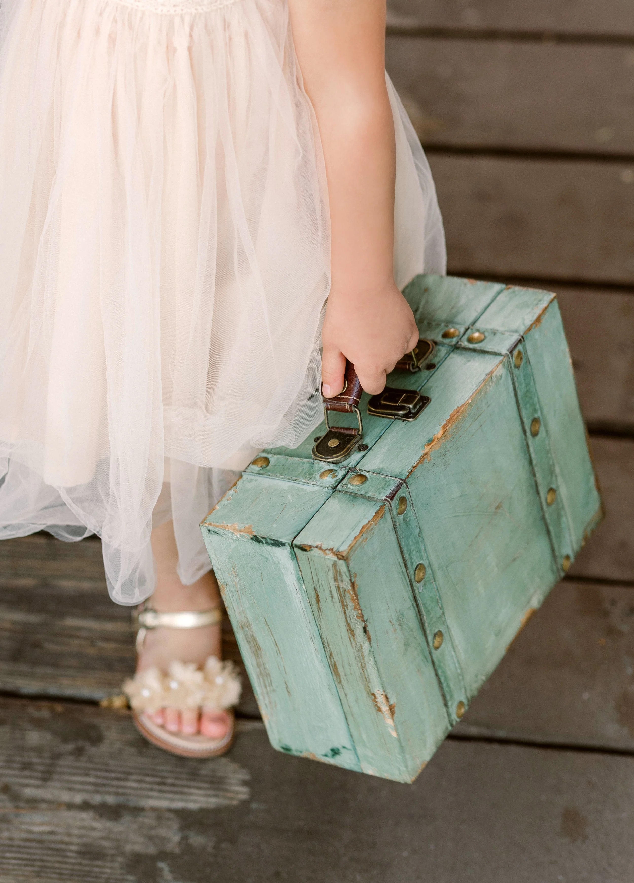A child's hand holding a vintage teal suitcase, wearing a pink tulle dress and flower-adorned sandals, standing on wooden planks.