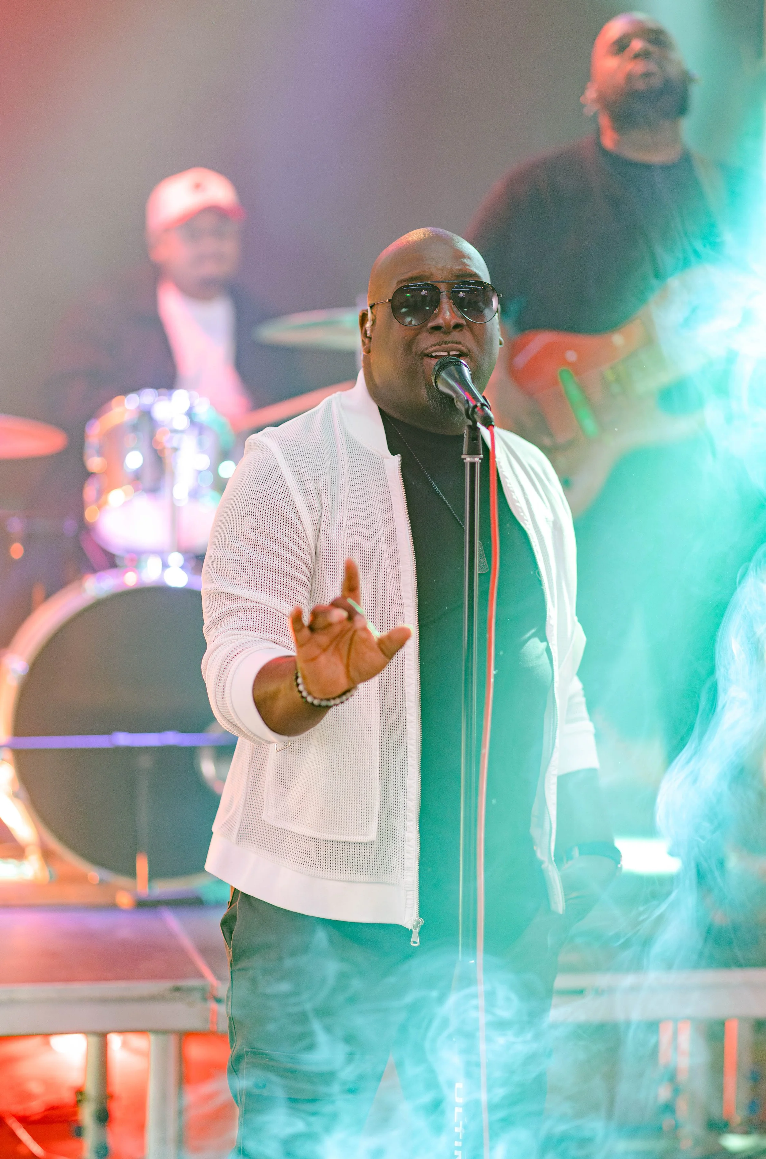 A man in sunglasses and a white jacket singing into a microphone on stage with a band playing instruments in the background, colorful stage lighting, and some green smoke.