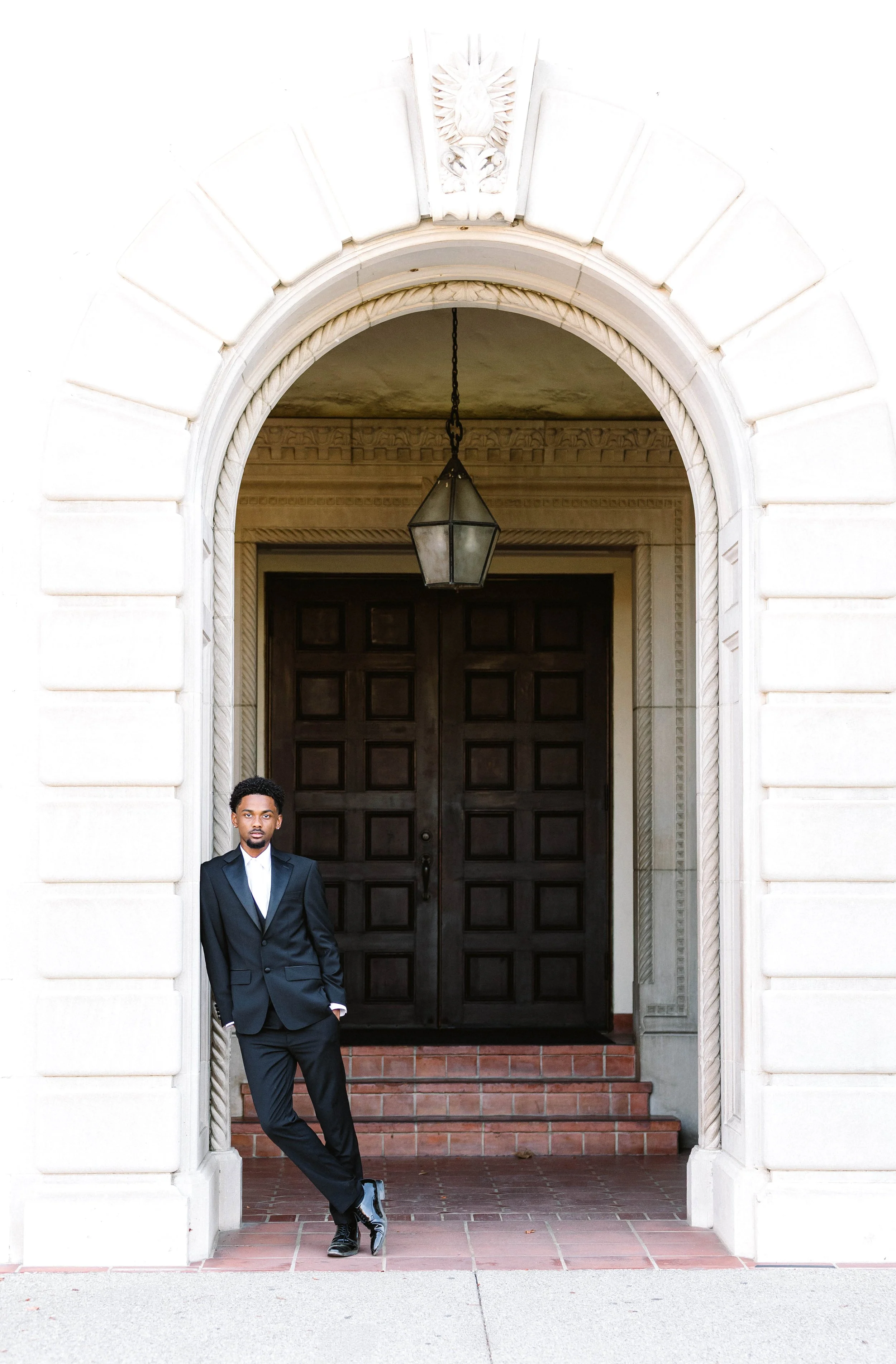 A man in a black tuxedo standing casually against a stone archway outside a building with a large dark wooden door and hanging lantern.