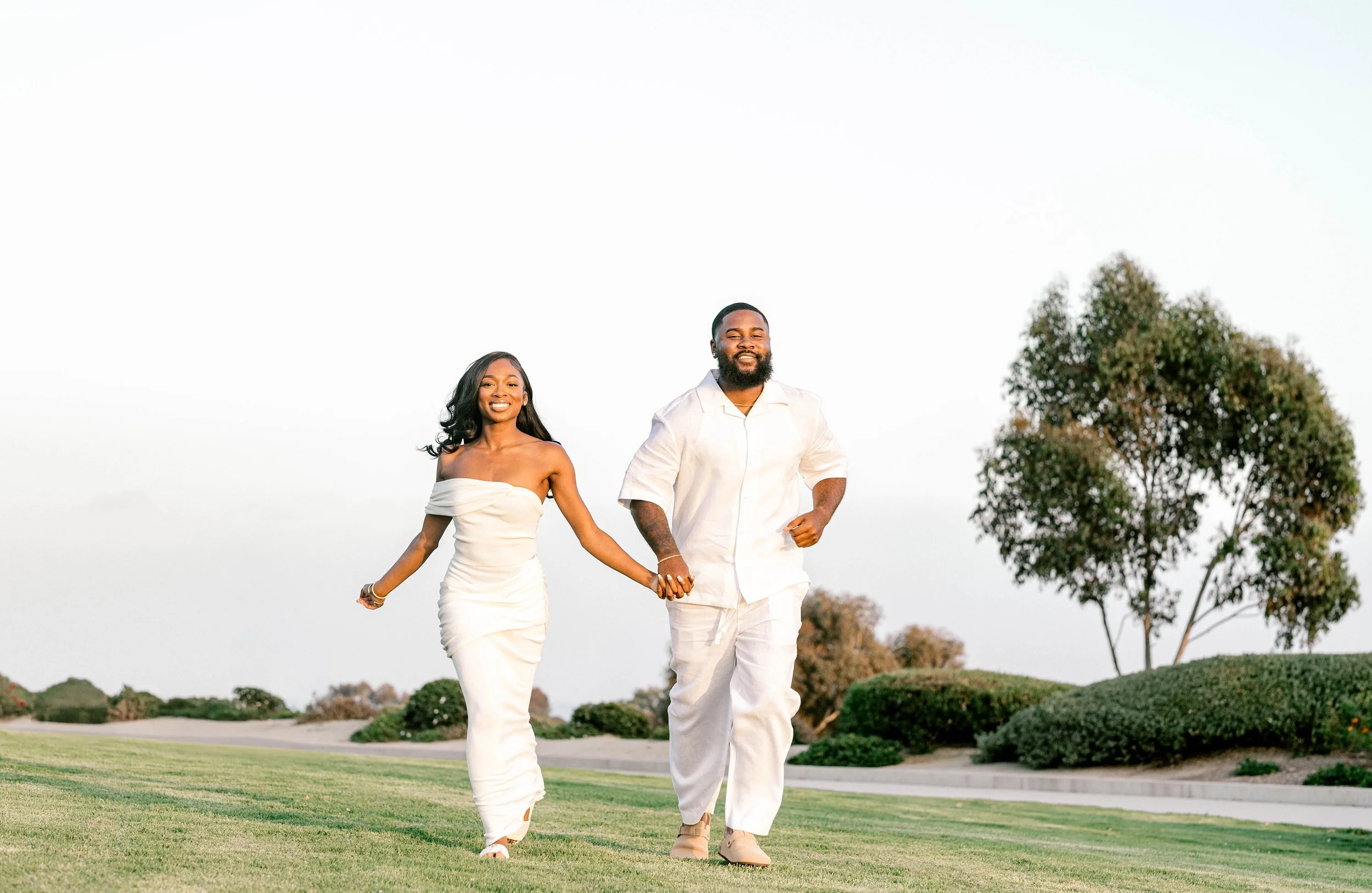 A smiling couple walking hand-in-hand on a grassy field, dressed in white, with trees and a clear sky in the background.