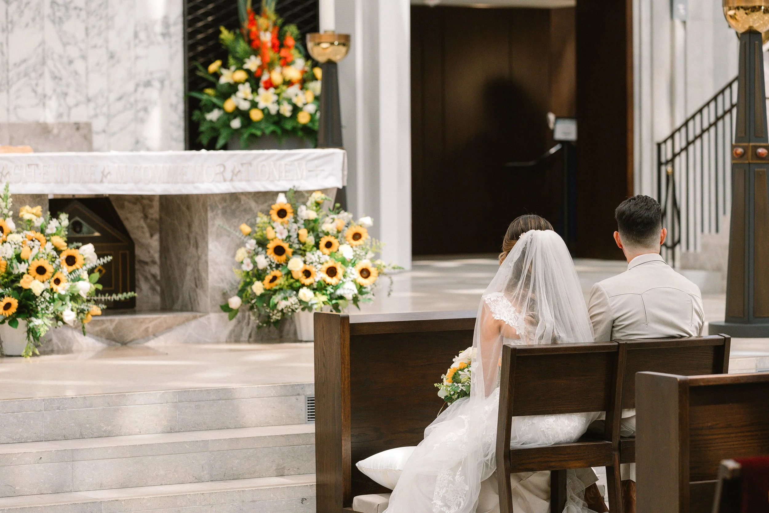 A bride and groom sitting on a wooden bench during a wedding ceremony inside a church, with floral arrangements and an altar in the background.