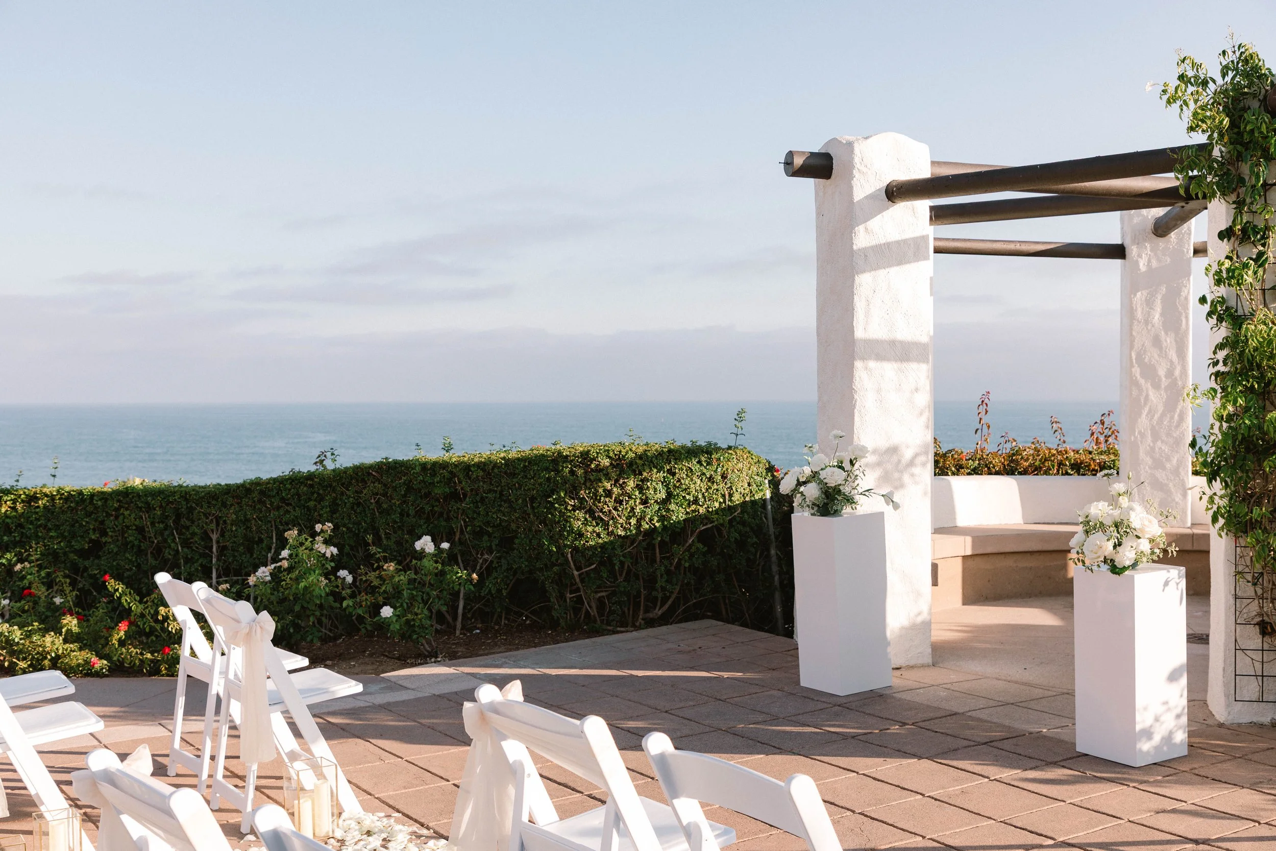 Outdoor wedding setup with white chairs decorated with ribbons, floral arrangements, and an altar with floral decor, overlooking the ocean with a clear sky.