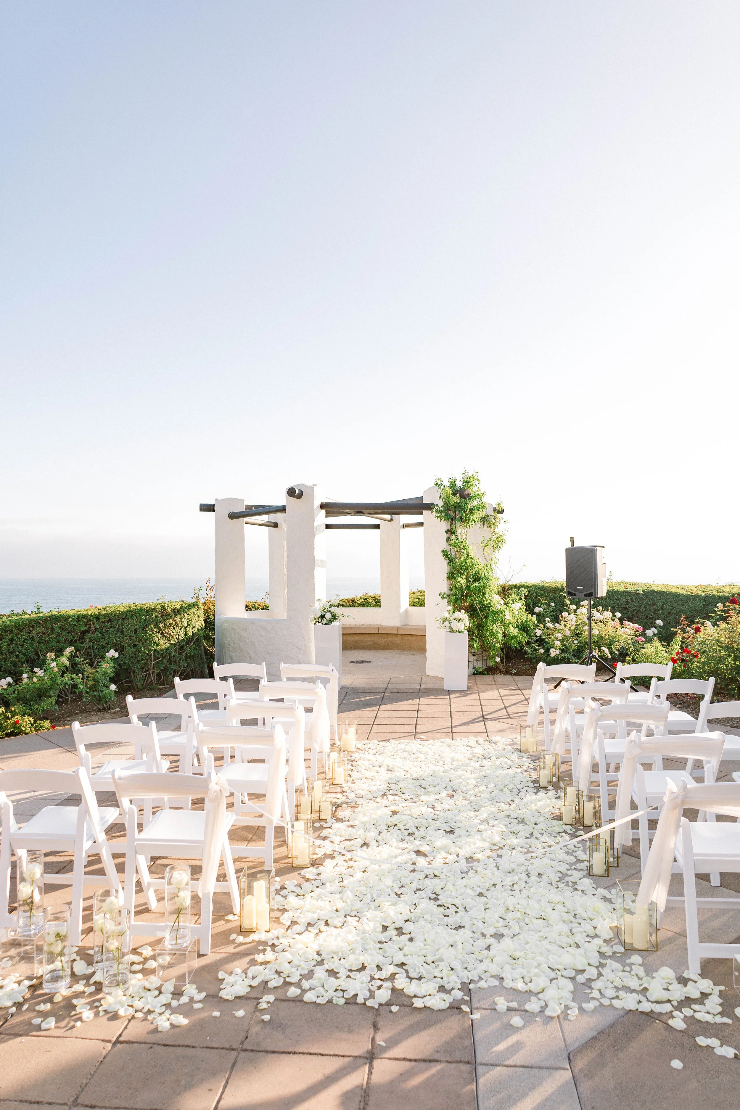 Outdoor wedding ceremony setup with white chairs, rose petal aisle, candles, and a small stage with a pergola in a garden overlooking the ocean.