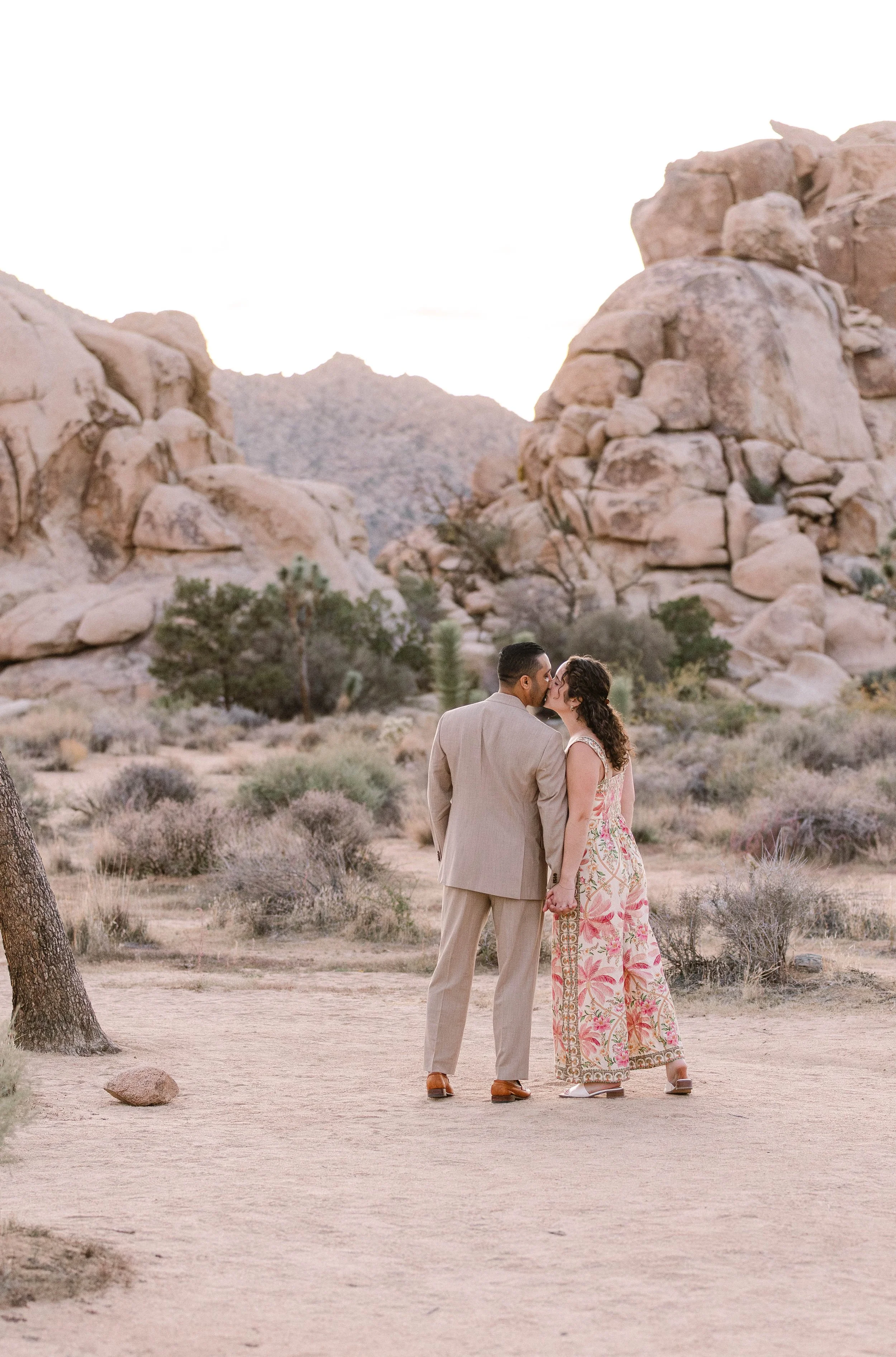 A couple holding hands and kissing in a desert landscape with large rock formations and bushes.
