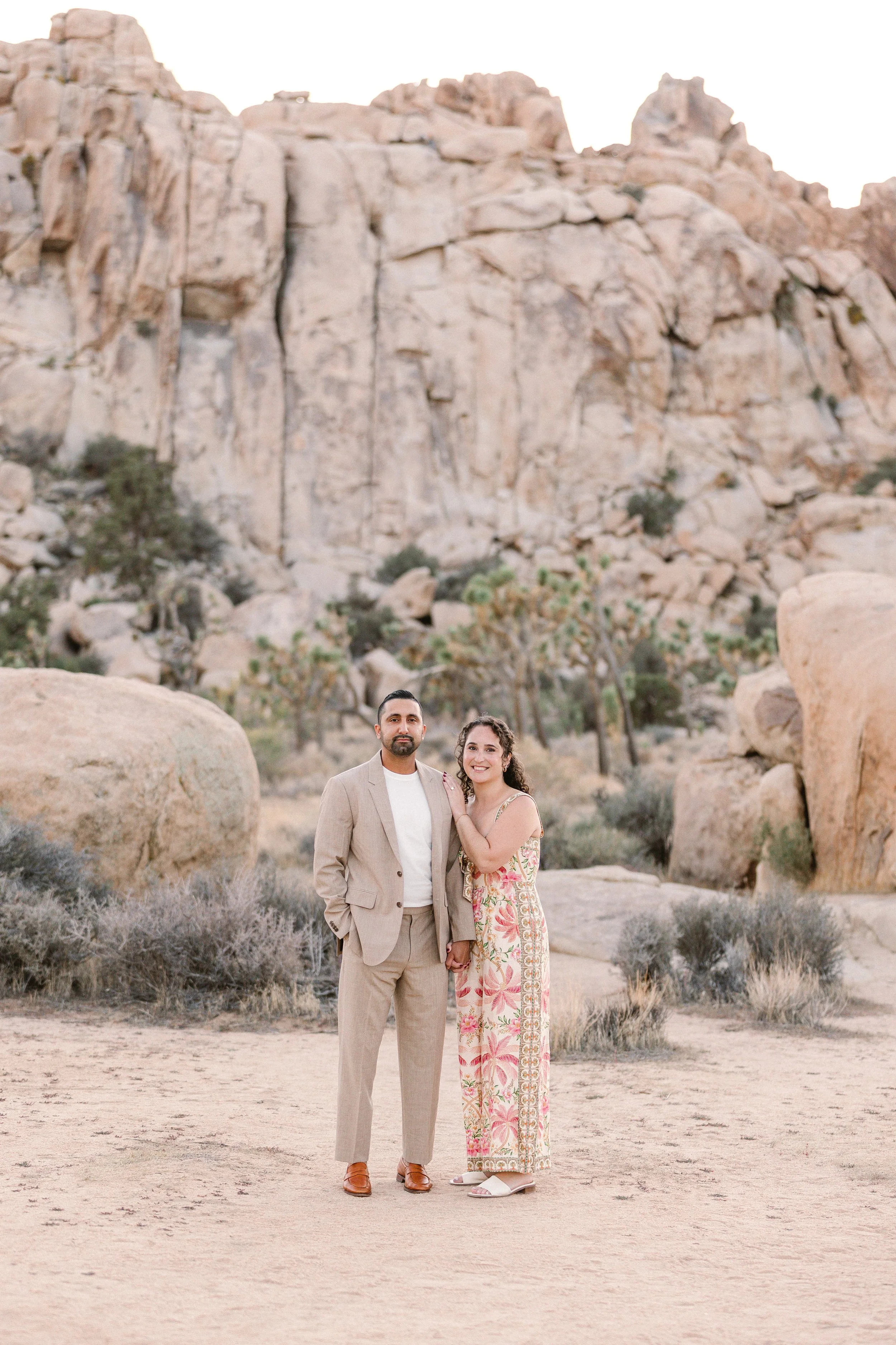 A couple stands hand in hand in a desert landscape with large rocks and desert plants, smiling at the camera.