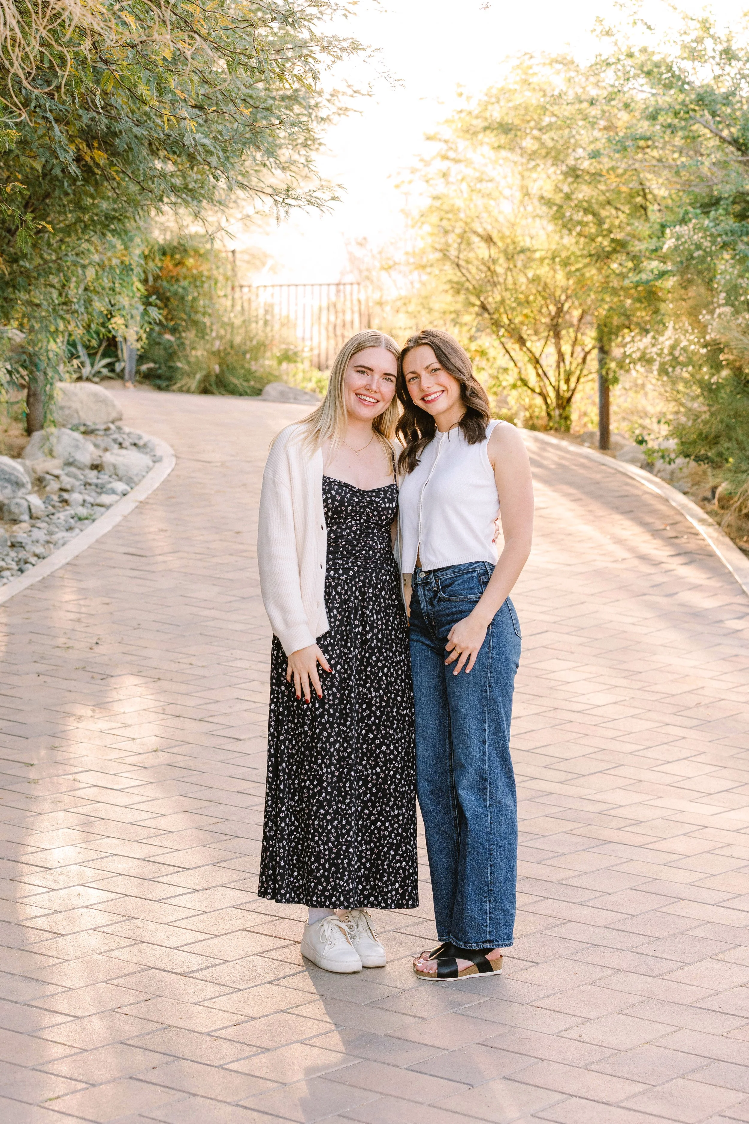 Two young women standing outdoors on a brick pathway, smiling at the camera with trees and sunlight in the background.