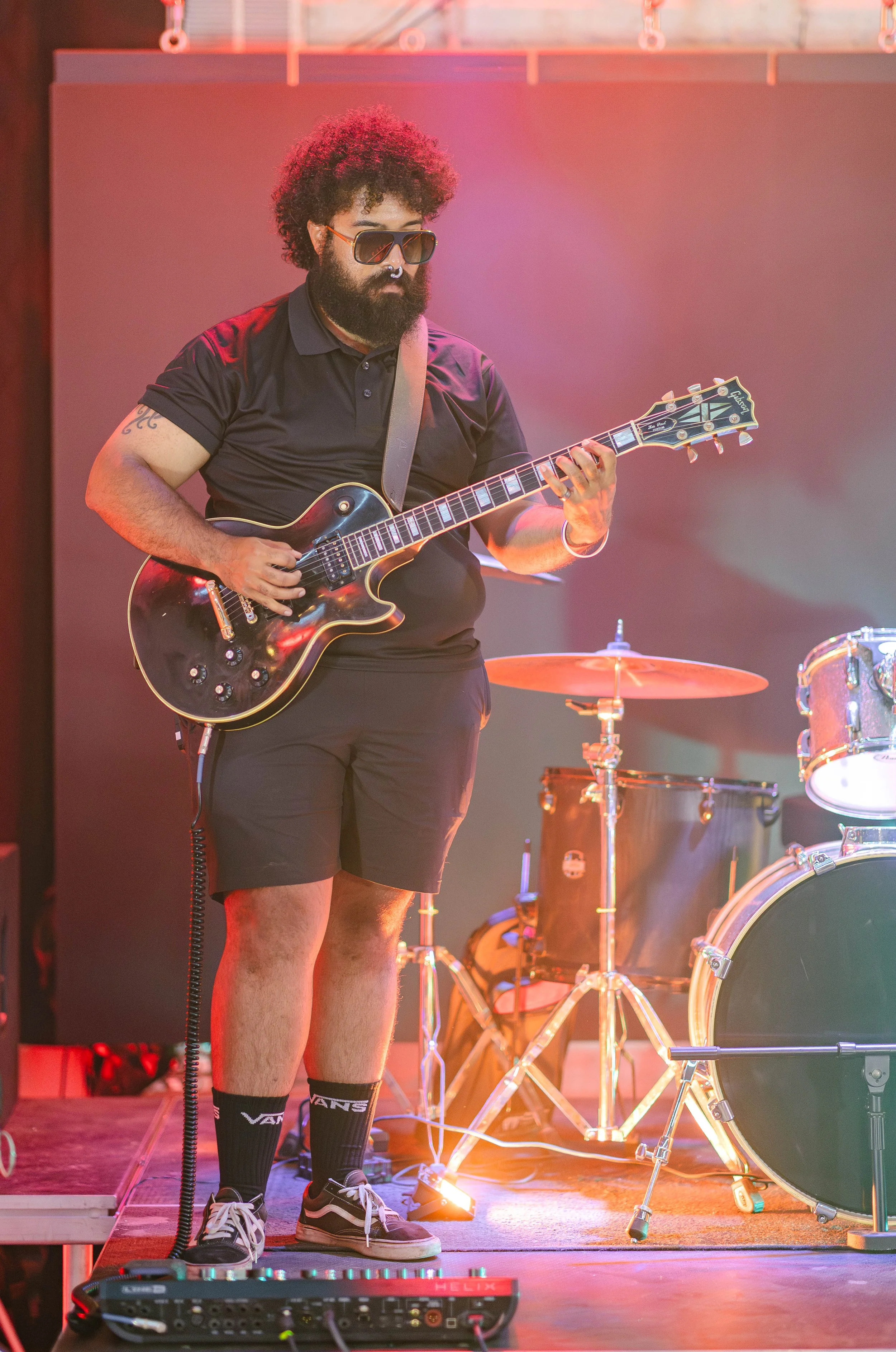 A man with curly hair, sunglasses, and a beard plays an electric guitar on stage, wearing a black polo shirt, shorts, and Vans socks.