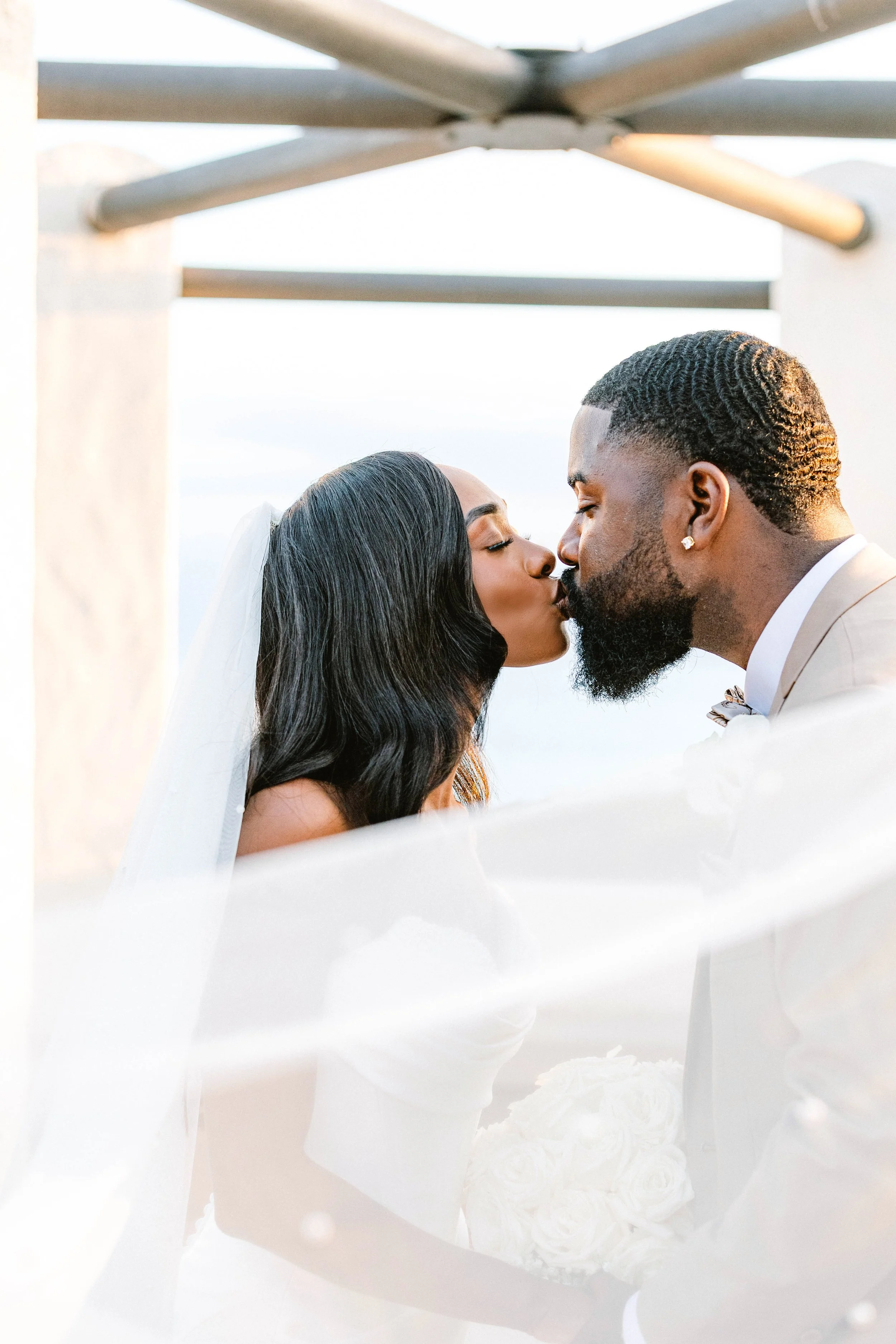 A newlywed couple sharing a kiss during their wedding ceremony, with the bride holding a bouquet of white roses, under a canopy with a soft natural light.