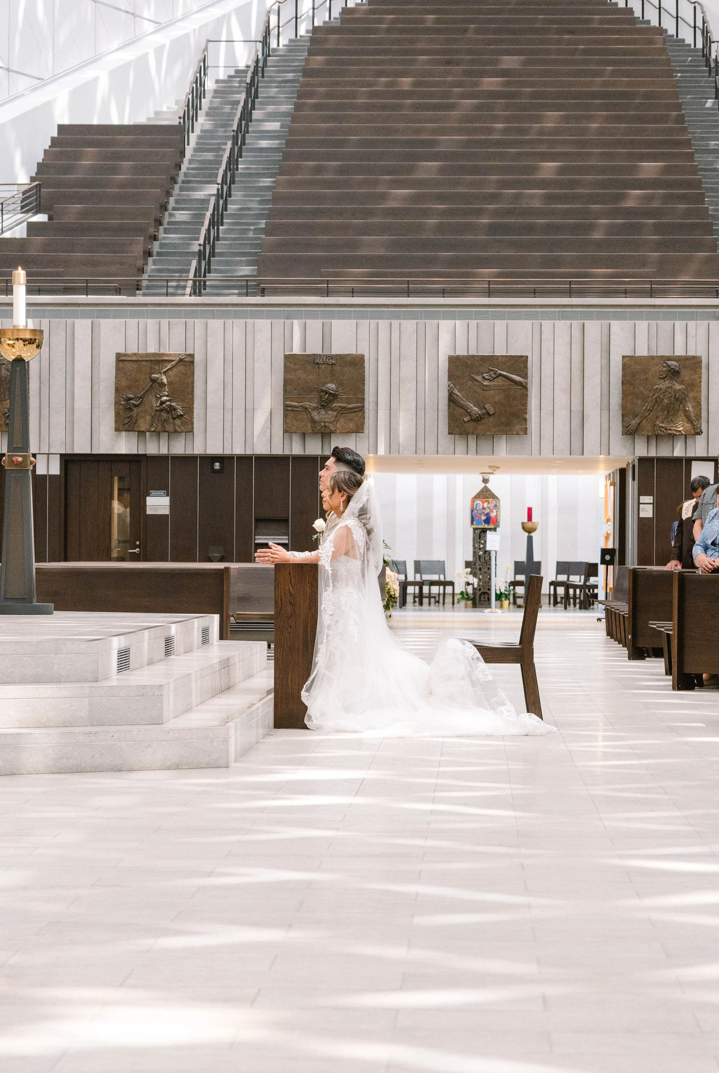 A bride kneeling at the altar during a wedding ceremony inside a church. She is wearing a white lace wedding dress and veil, with her hands together. The church has a large, open space with pews, sculptures on the wall, a large staircase in the backg
