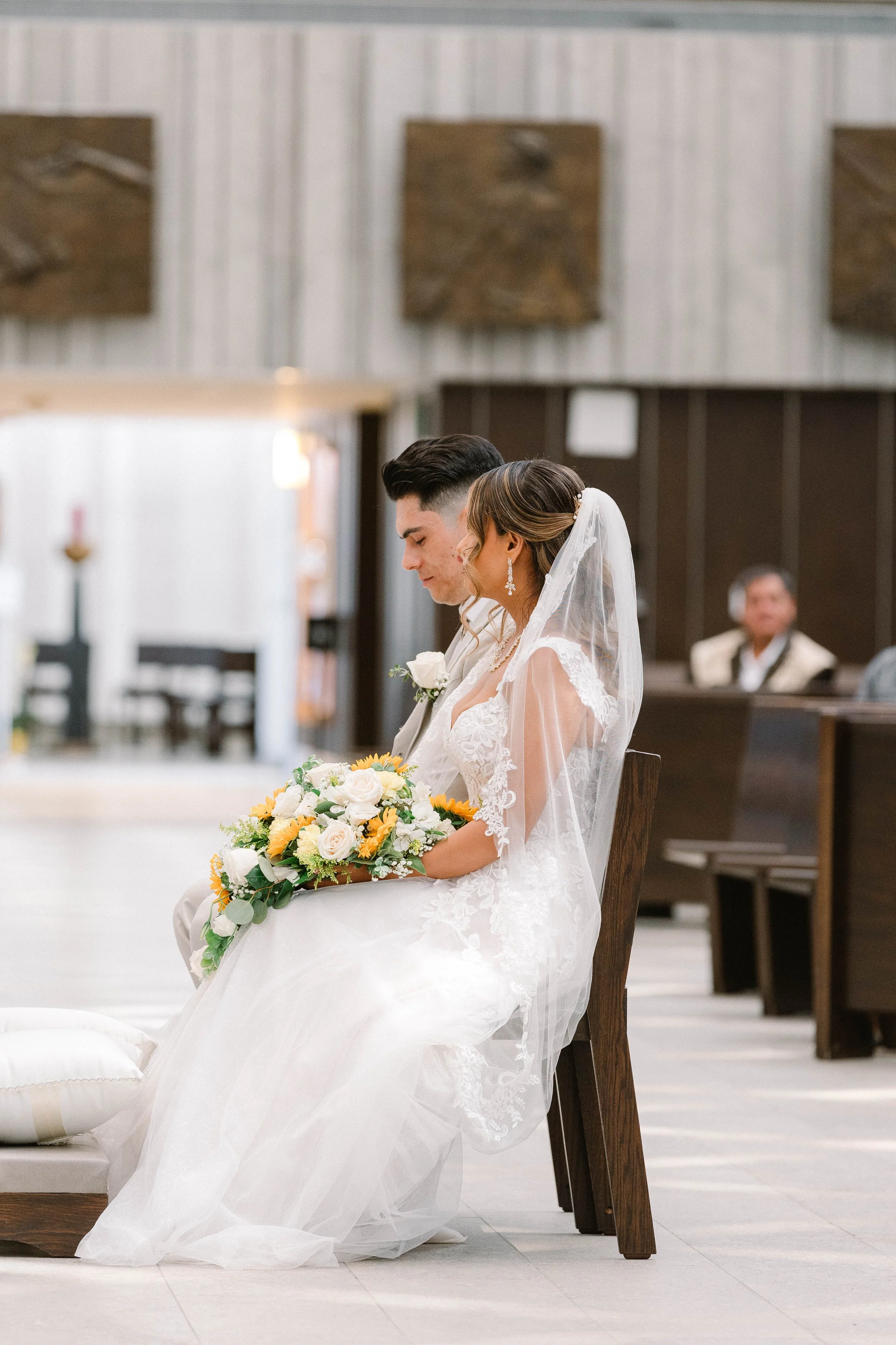 A bride and groom sitting in a church during their wedding ceremony, with the bride holding a bouquet of white and yellow flowers, dressed in white lace, wearing a veil, and the groom in a beige suit with a white shirt and boutonniere.