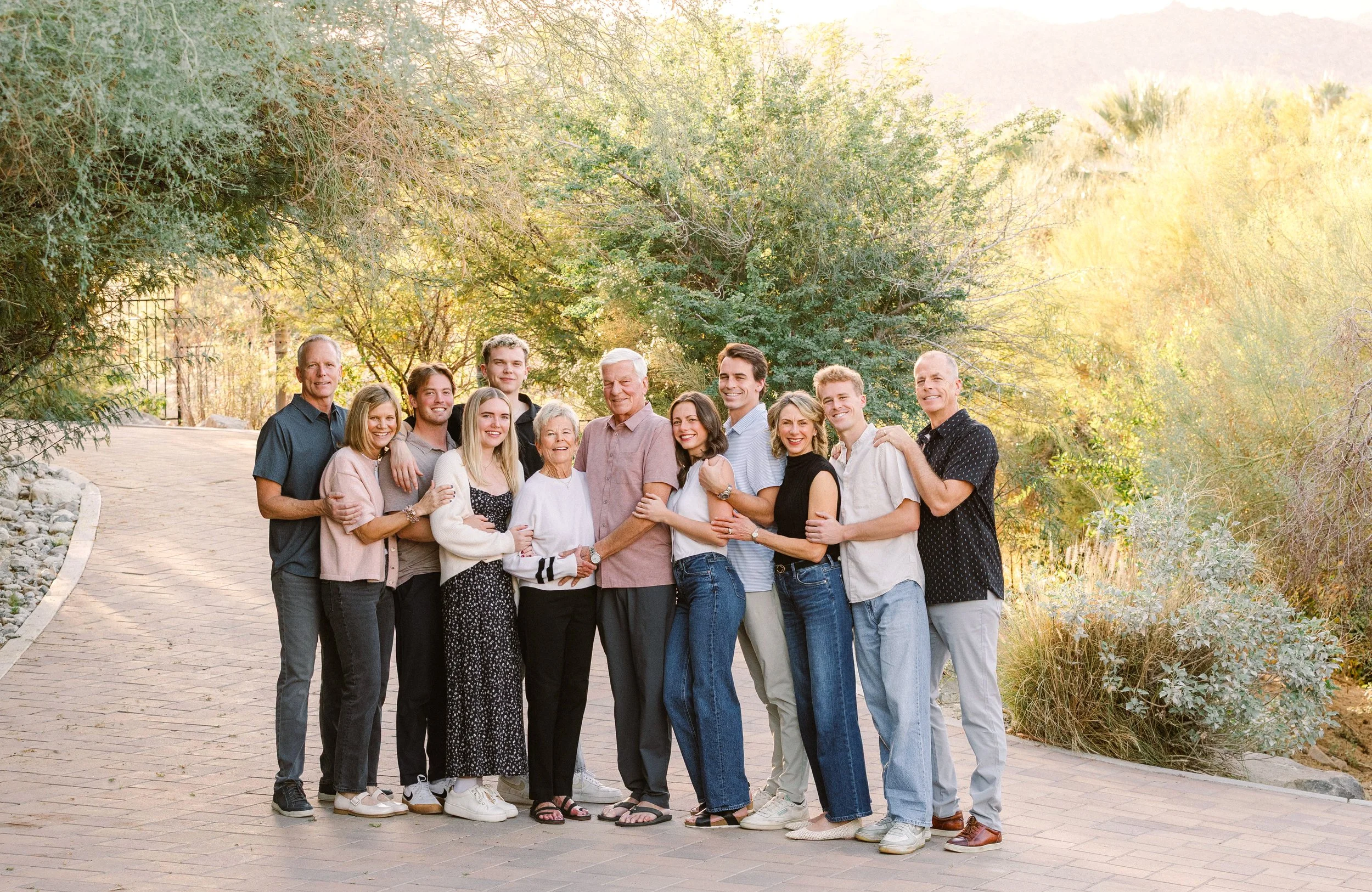 A large, multigenerational family standing outdoors on a paved pathway surrounded by trees and greenery, smiling and embracing each other.