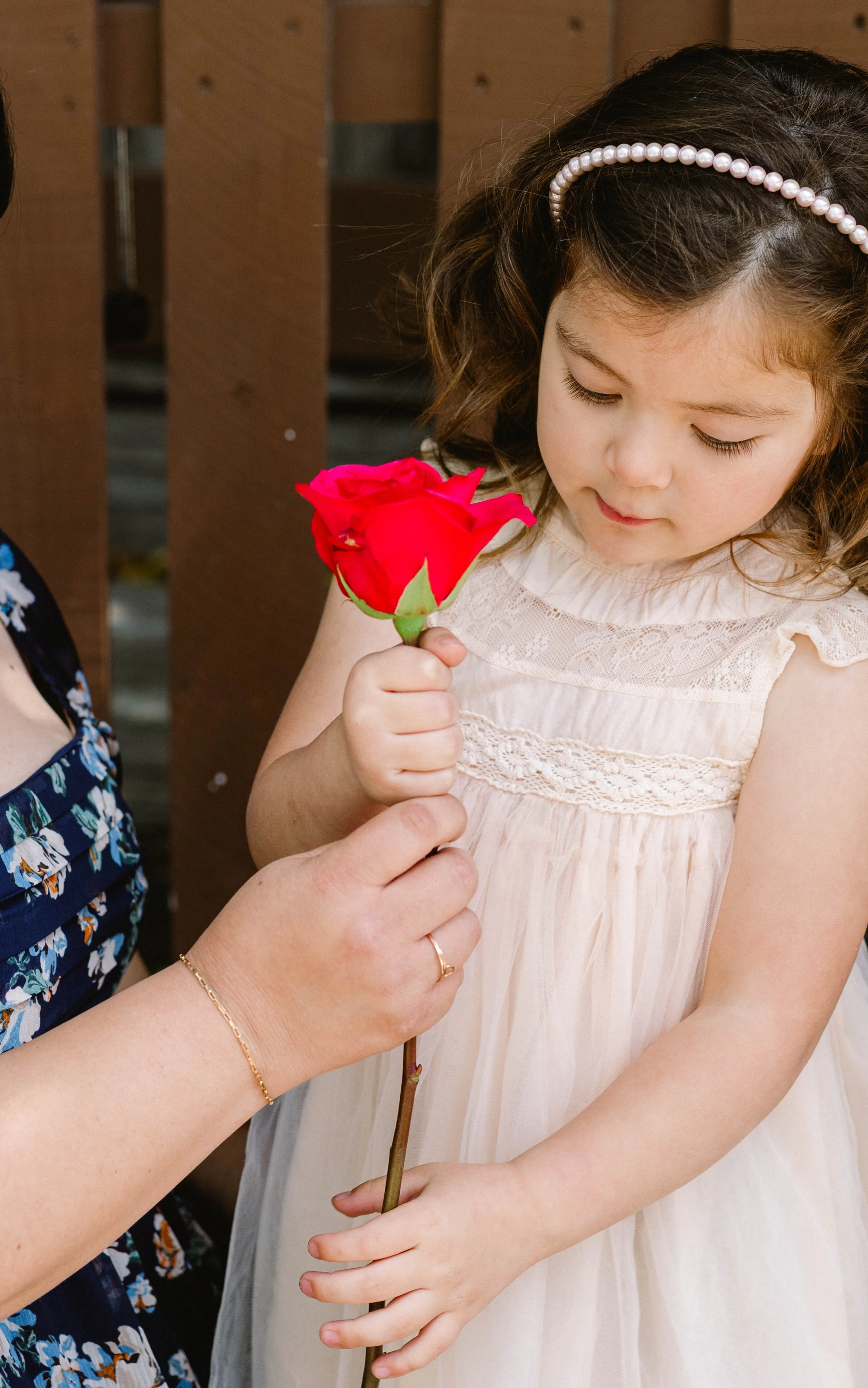 A young girl in a cream-colored dress with a pearl headband is being handed a red rose by an adult with a gold bracelet and wedding ring.