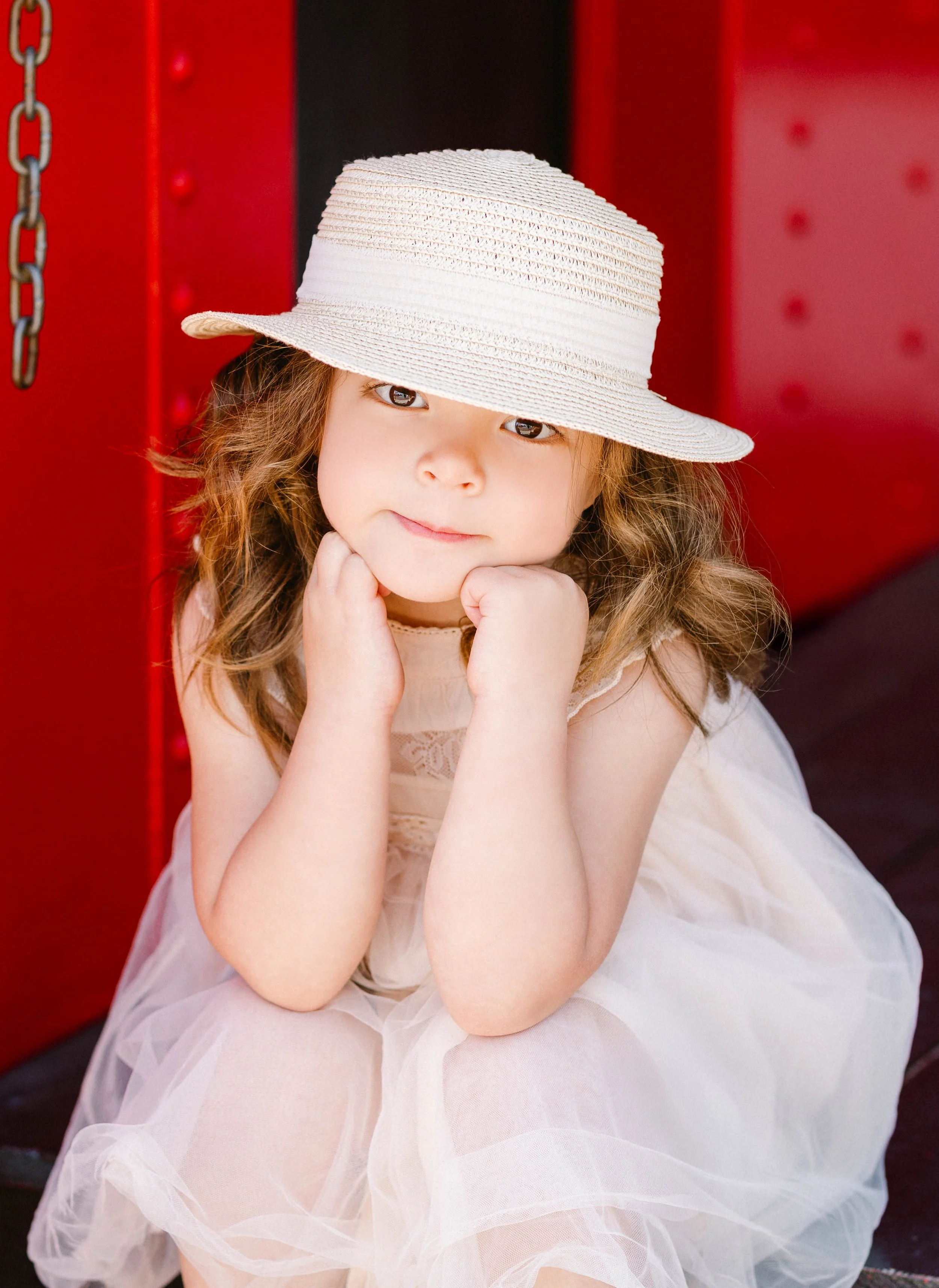 A young girl with curly hair, wearing a cream-colored dress and a wide-brimmed straw hat, resting her chin on her hands with a thoughtful expression, seated against a red background.