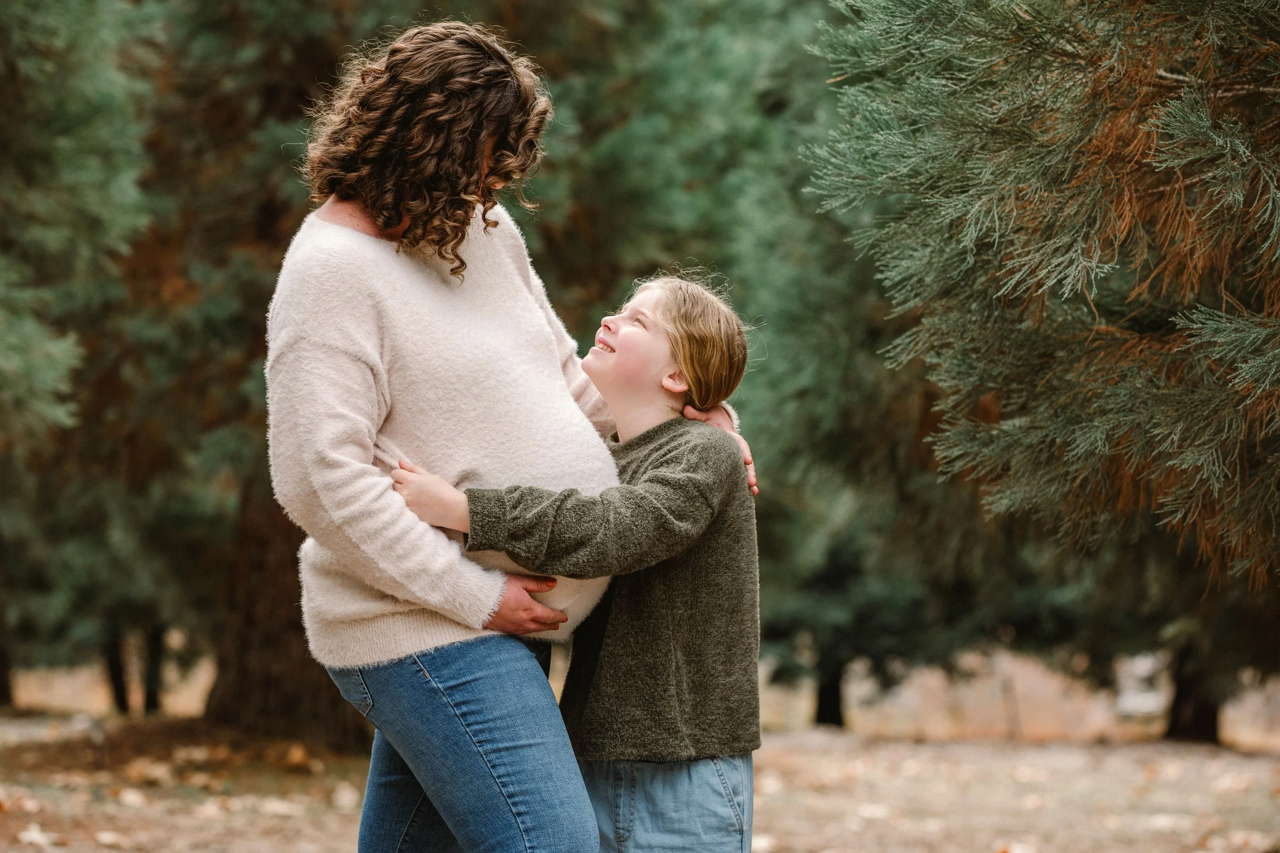 A woman and a young girl sharing a joyful moment outdoors among evergreen trees, with the woman holding the girl's hands and both smiling.