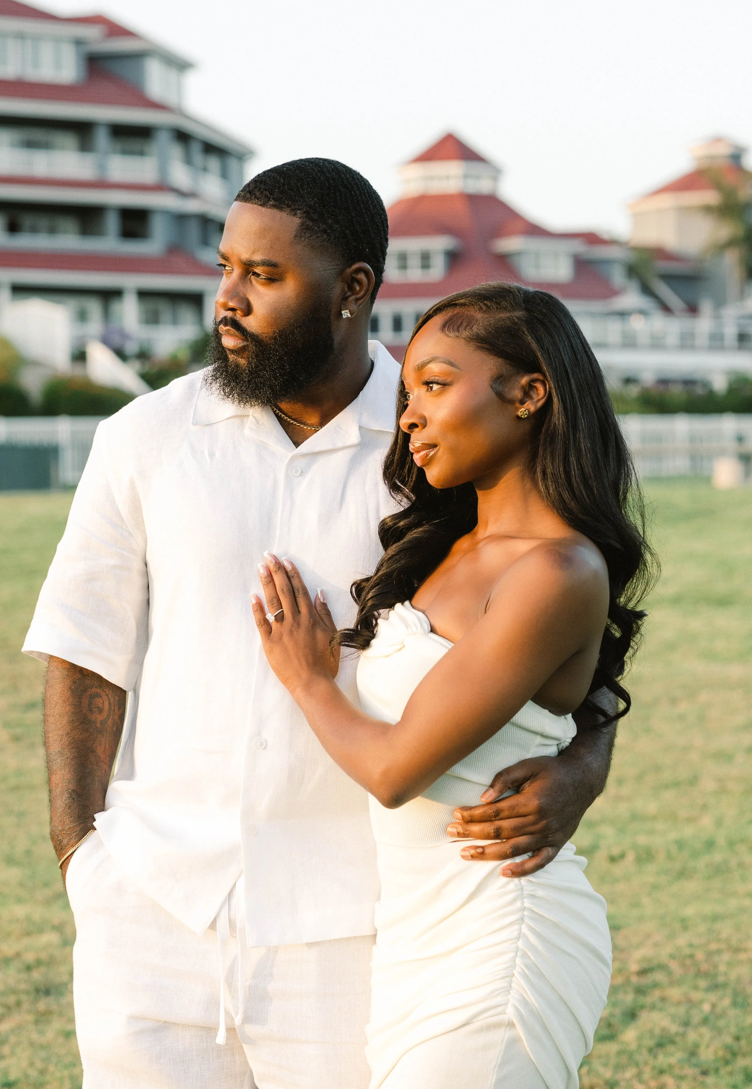 A couple standing outdoors on a lawn, with the man in a white shirt and the woman in a white dress, looking at each other.