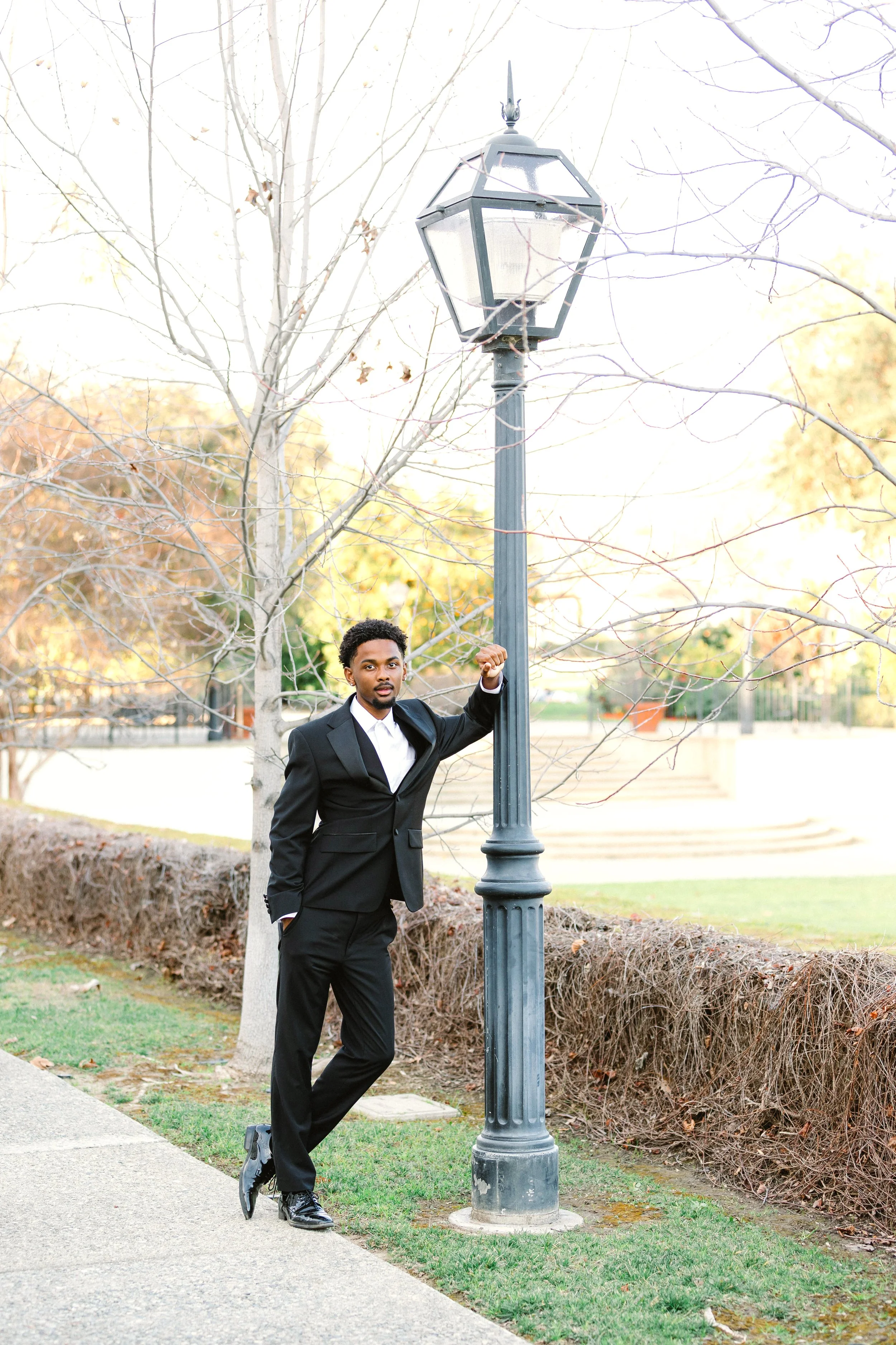 A man in a black suit and white shirt standing outdoors beside a tall black lamppost next to leafless trees.