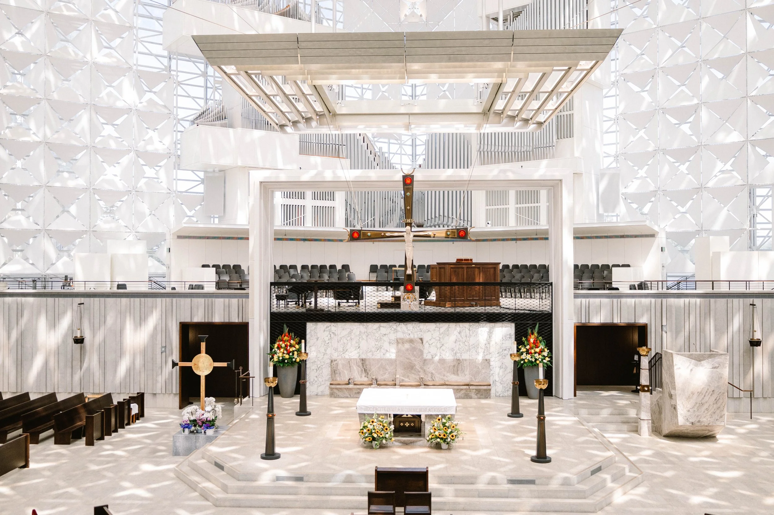 Interior of a modern church with an altar, crucifix, and floral arrangements, featuring unique architectural design with bright lighting and white geometric walls.