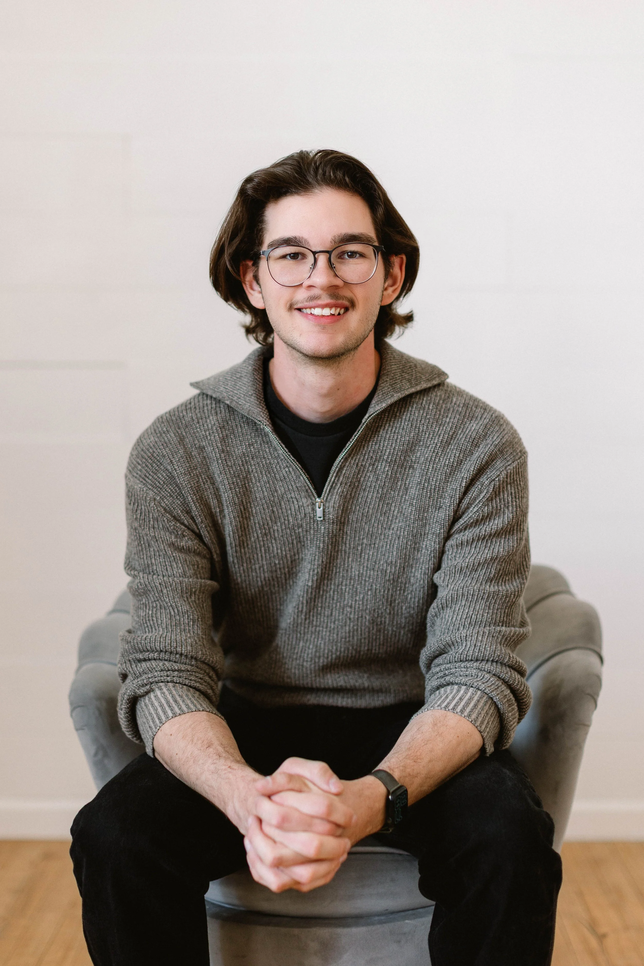A young man with glasses and brown hair, dressed in a gray zip-up sweater, sitting on a light gray armchair against a white wall, smiling at the camera.