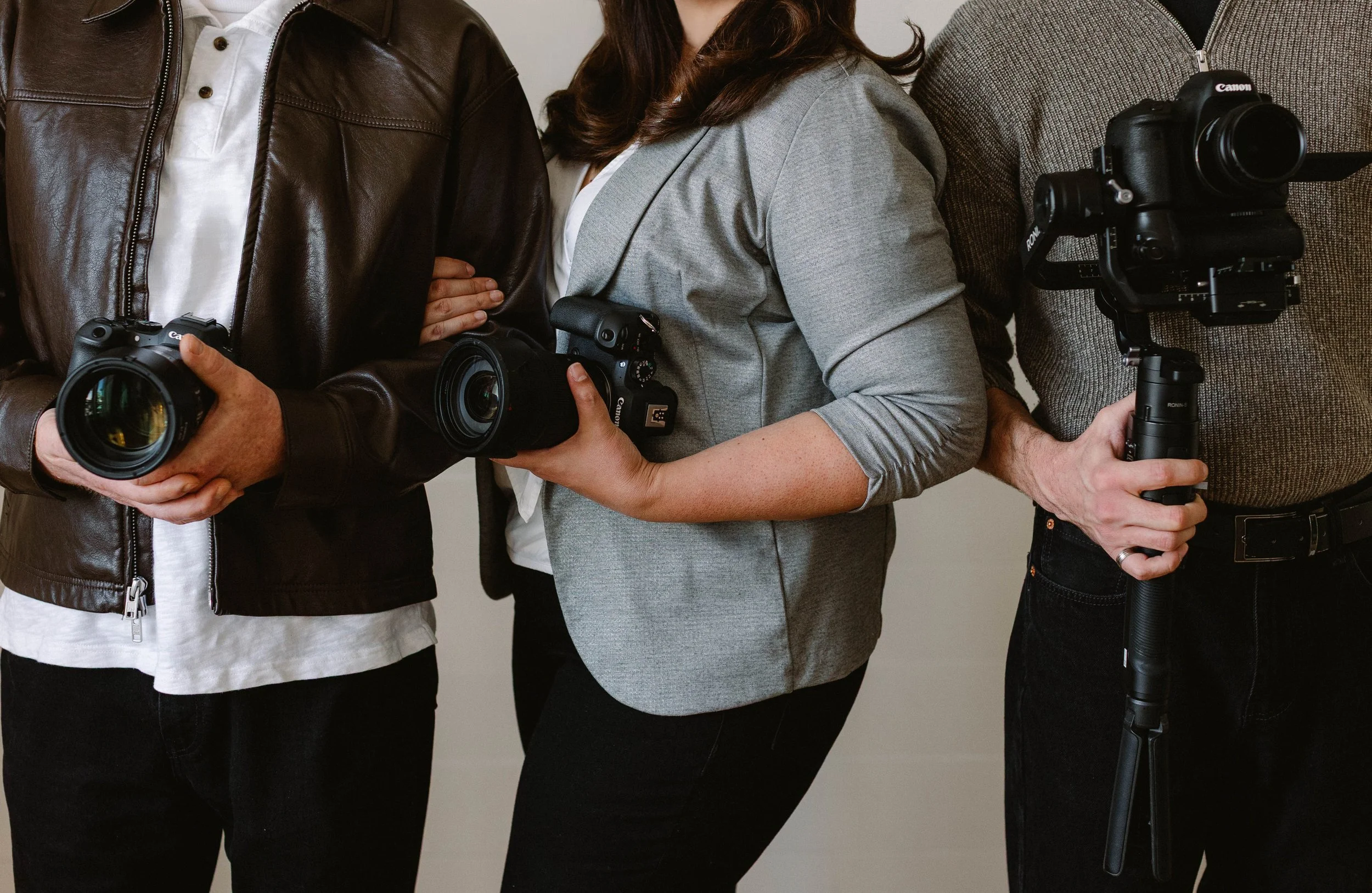 Three people standing indoors, each holding a camera. The person on the left is wearing a brown leather jacket, the person in the middle is wearing a gray blazer, and the person on the right is wearing a gray sweater.