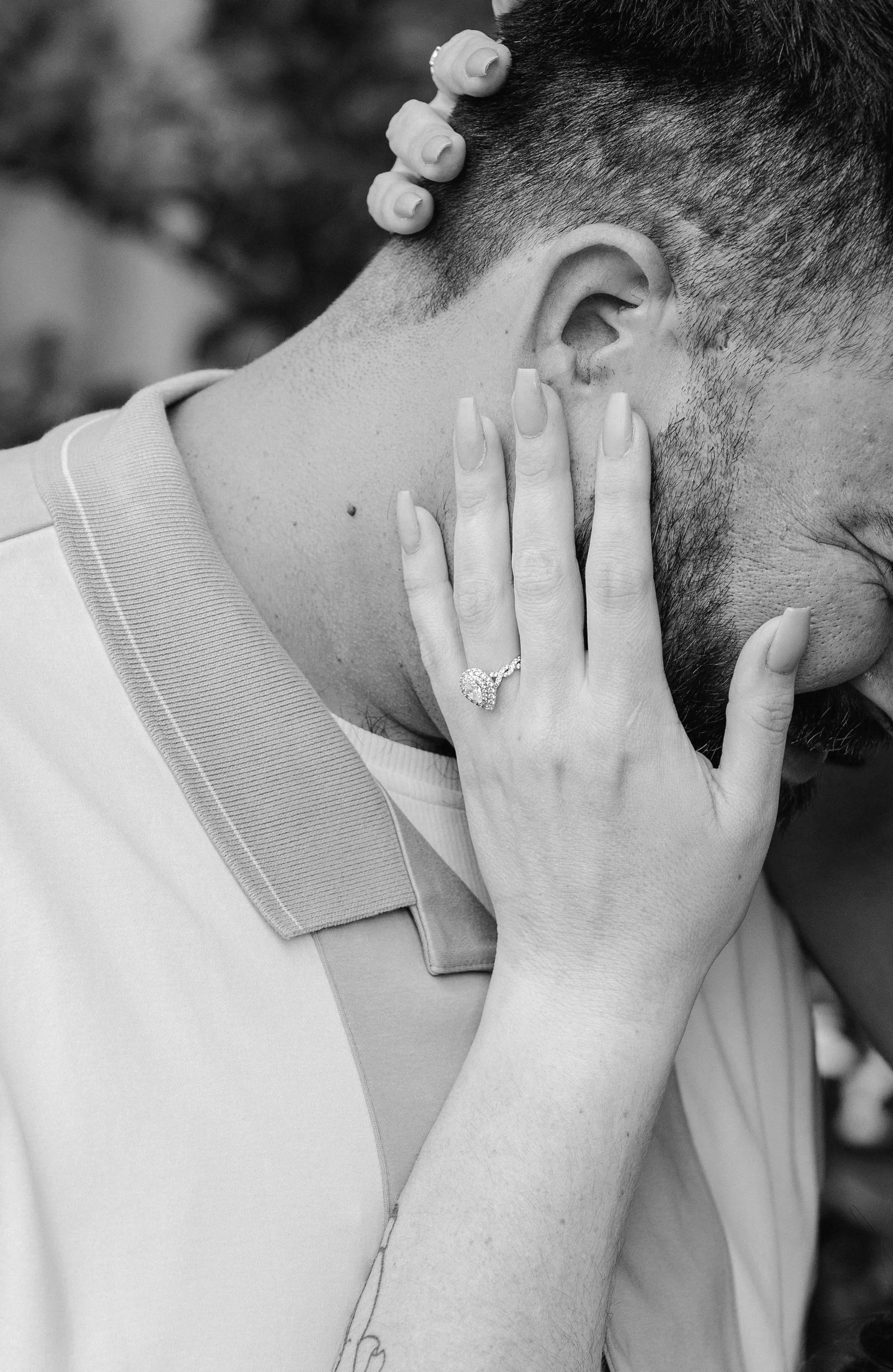 A person with a groomed beard and short hair touching their ear with a woman's hand adorned with a diamond ring and long nails resting on their neck.