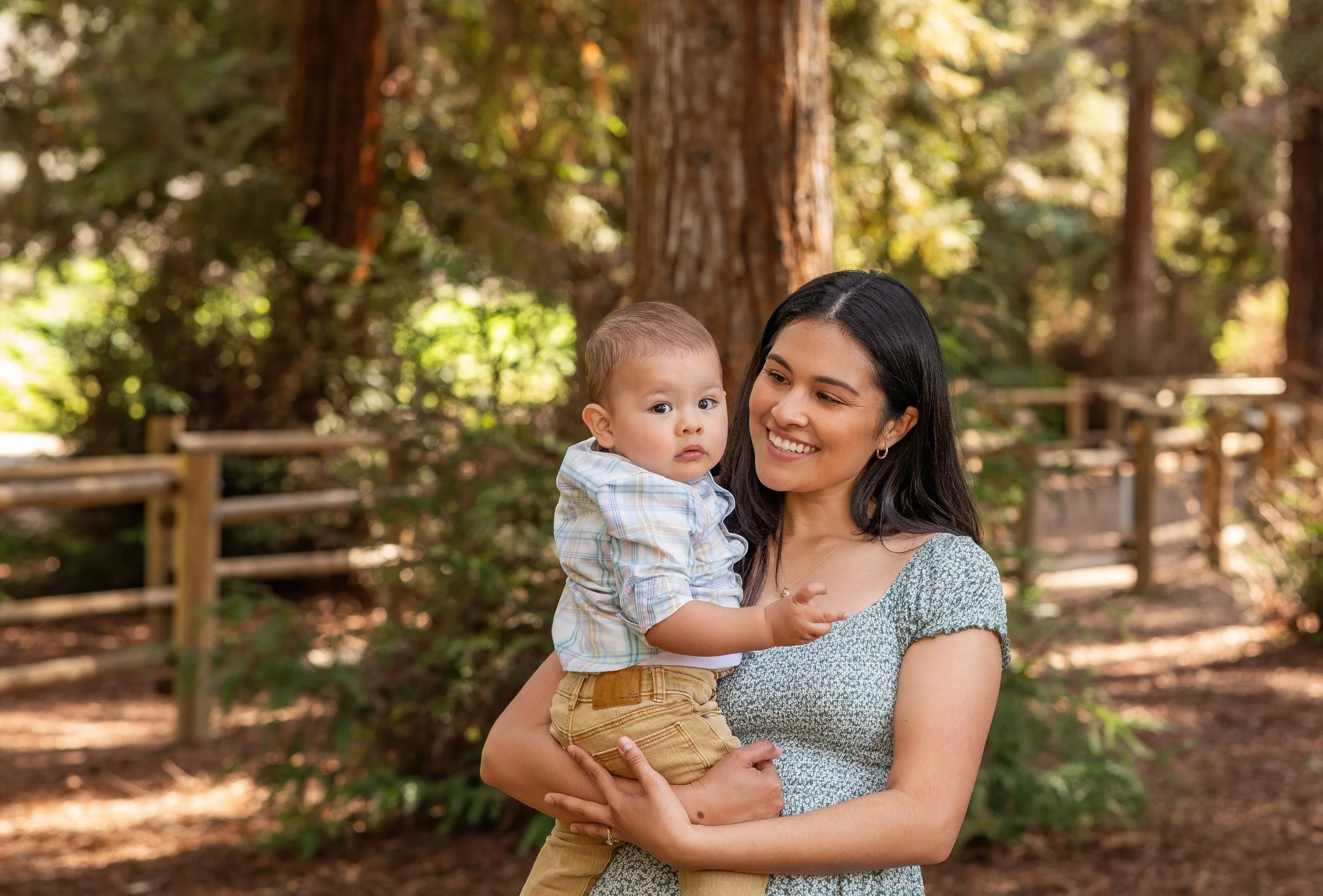 A smiling woman holding a young child outdoors in a wooded area with trees and a fence in the background.
