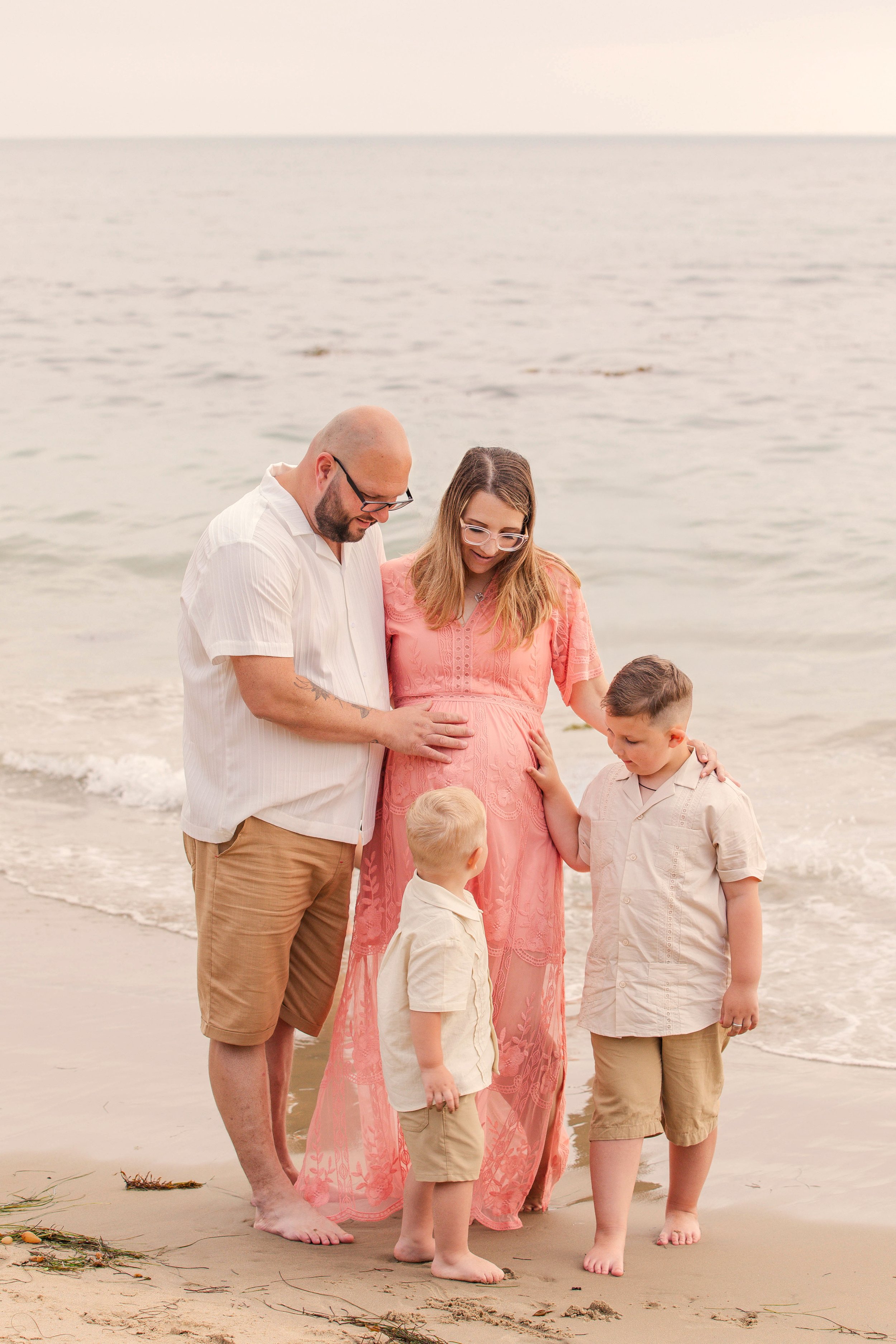 Family of four standing on a beach, with a pregnant woman wearing a pink dress, a man touching her belly, and two young boys in matching outfits.
