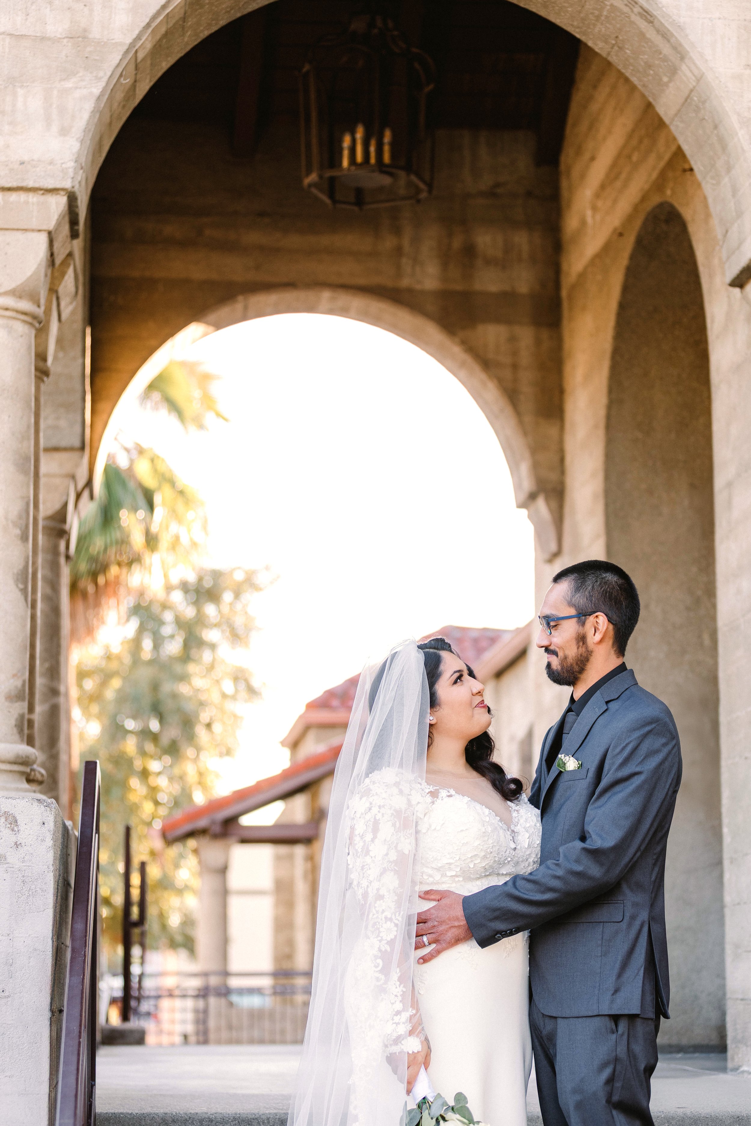A bride and groom standing close to each other under a stone archway, gazing into each other's eyes during their wedding. The bride is wearing a white lace wedding dress and veil, and the groom is in a gray suit with glasses. The background shows a b