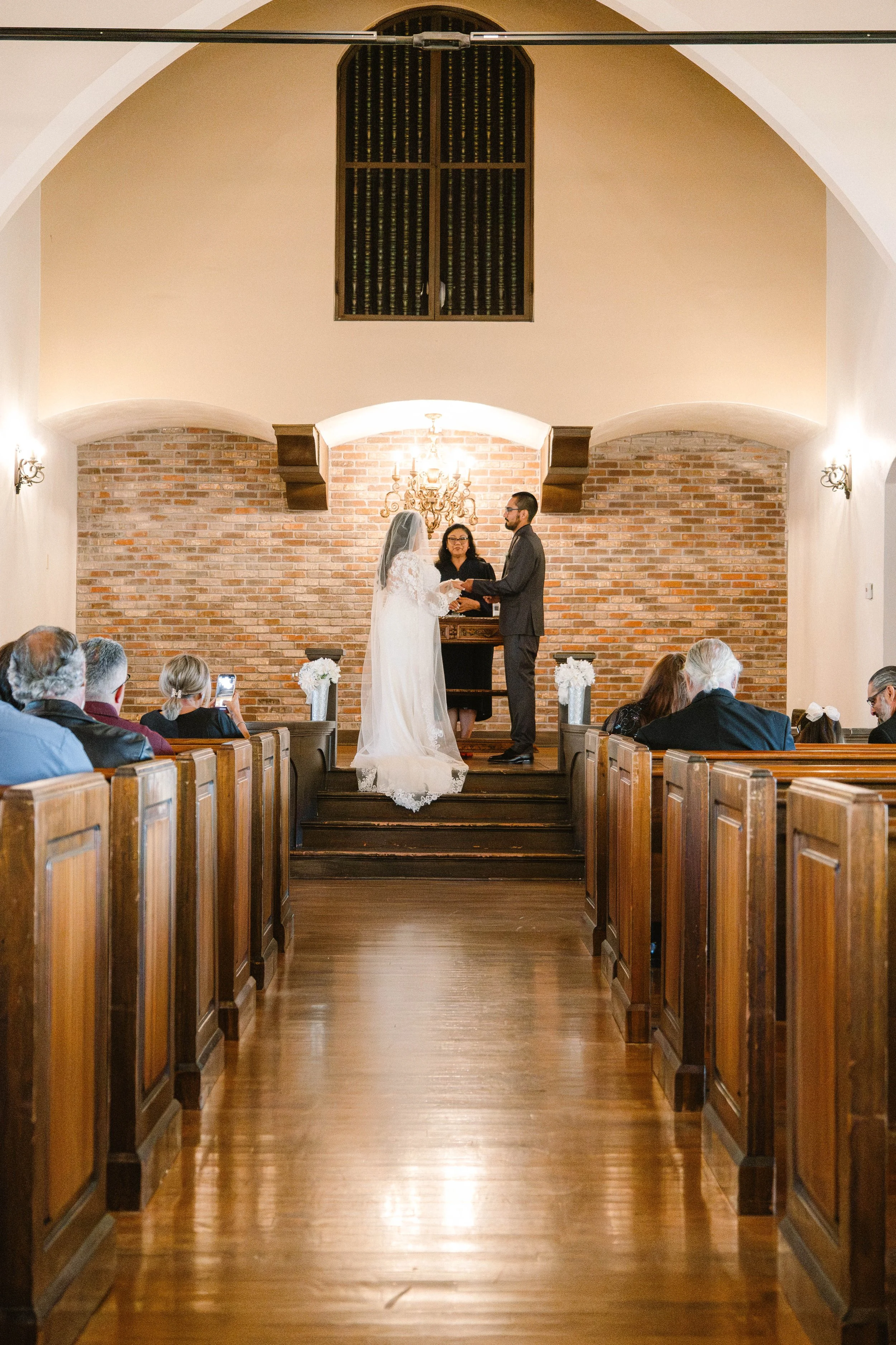 A bride and groom standing at the altar during their wedding ceremony inside a church, with guests seated in pews watching.