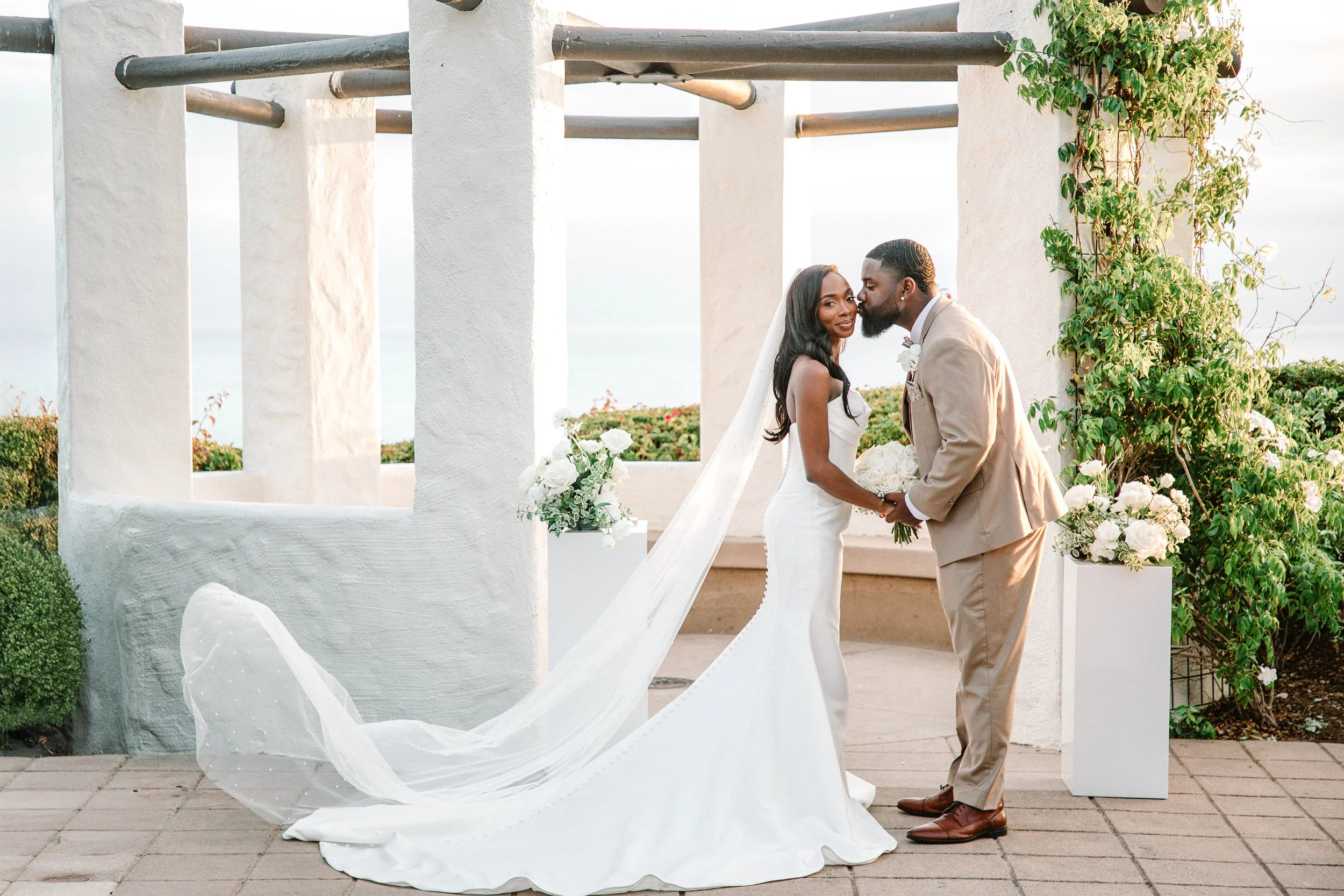 A newlywed couple holding hands and sharing a kiss at their outdoor wedding ceremony. The bride is wearing a white wedding gown with a long veil, and the groom is dressed in a beige suit. They are standing beneath a white architectural structure deco