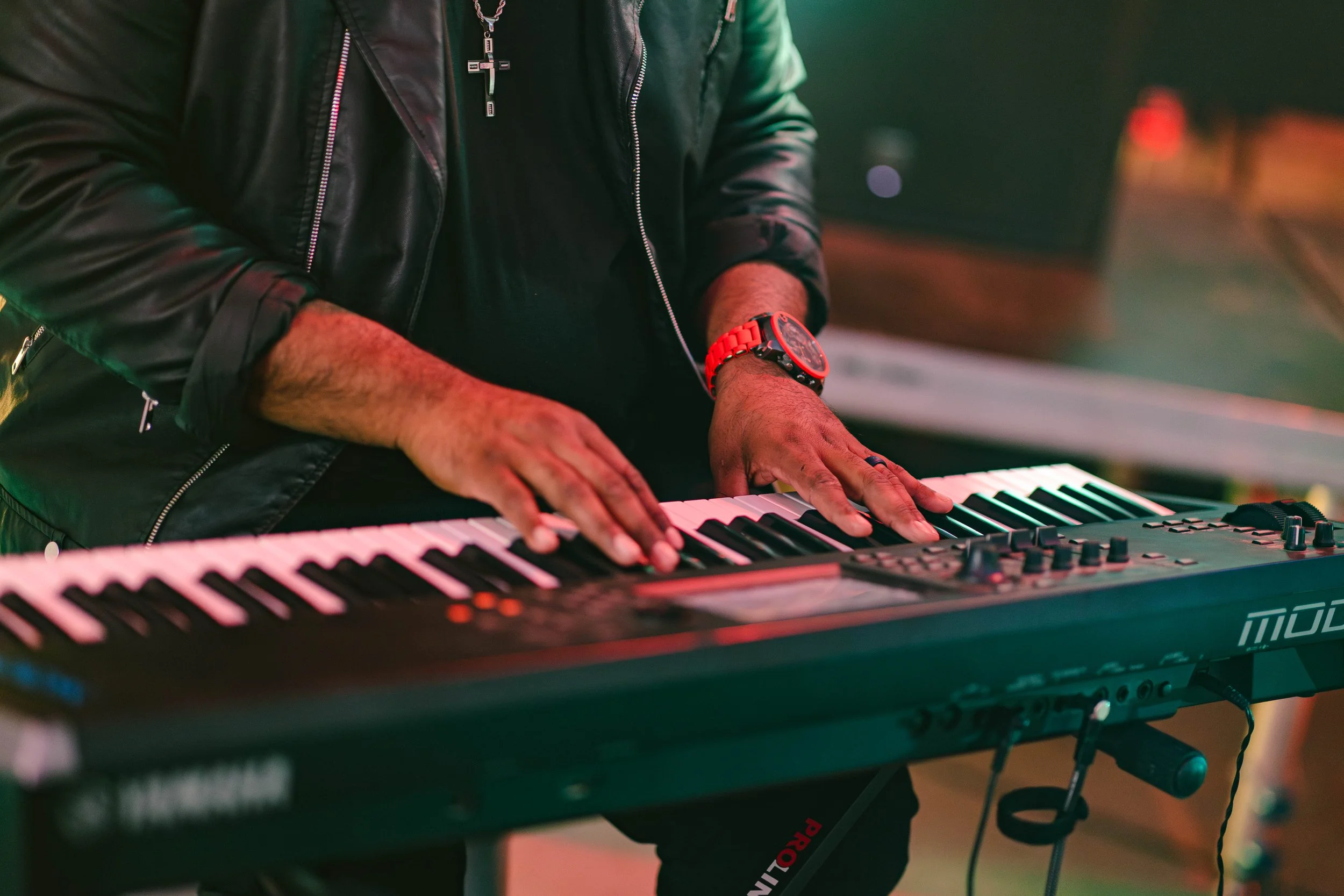 Person playing a keyboard on stage, wearing a black leather jacket, a large cross necklace, a red wristwatch, and rings, with a colorful, dimly lit background.