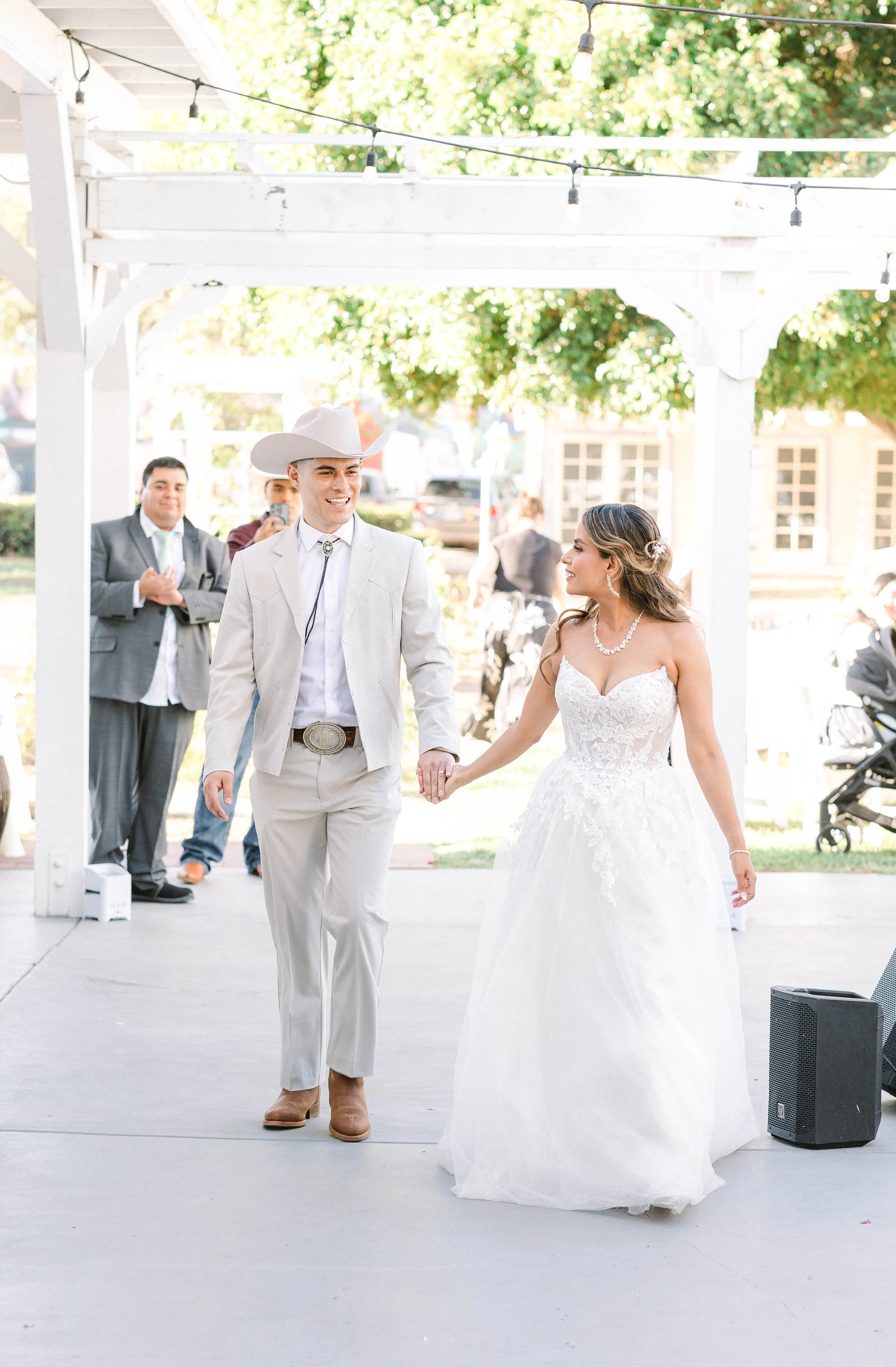Bride and groom holding hands during their wedding ceremony under outdoor white wooden arch with string lights.
