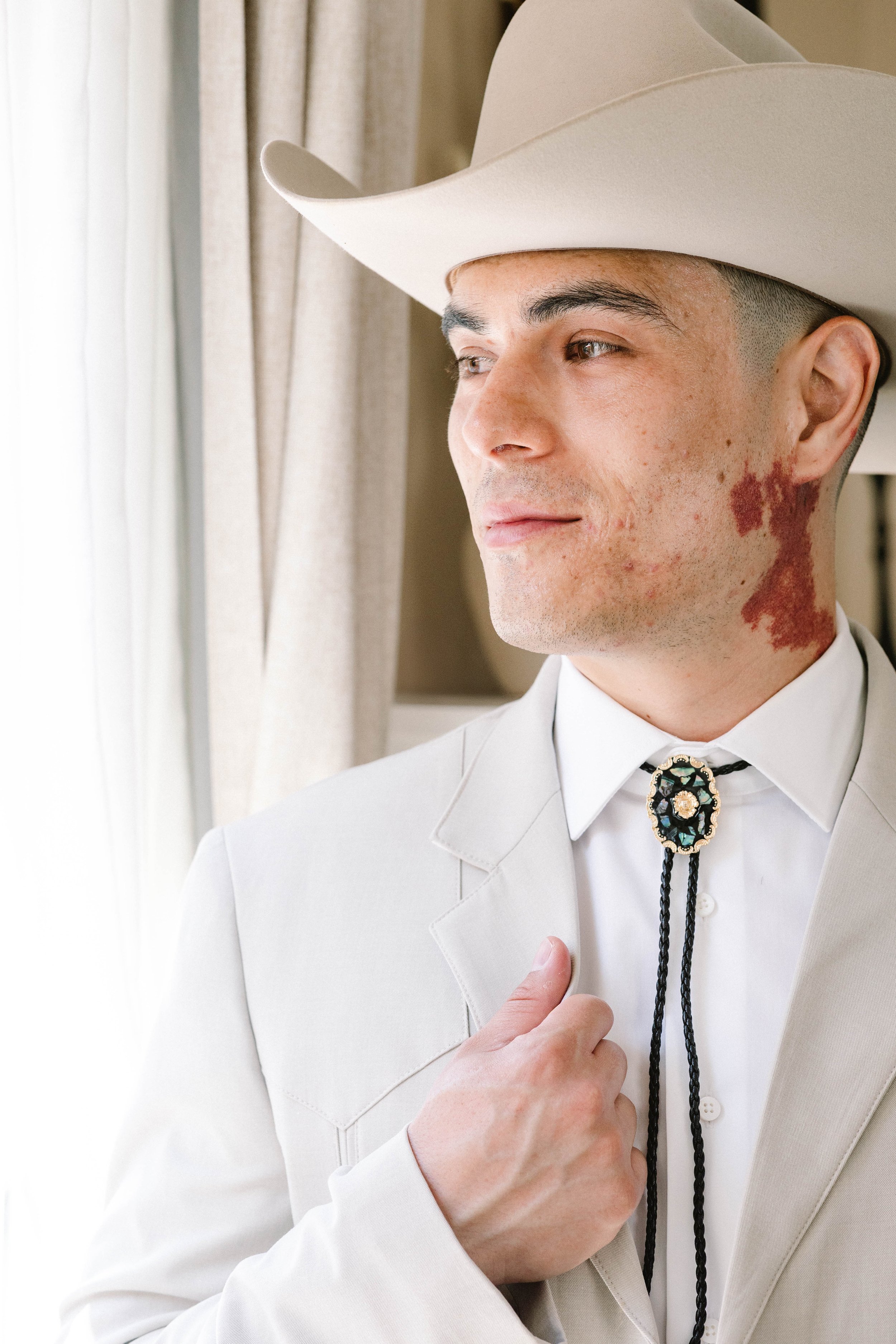 A man dressed in Western wear, wearing a cowboy hat, a cream suit, and a bolo tie, standing near a window with curtains.