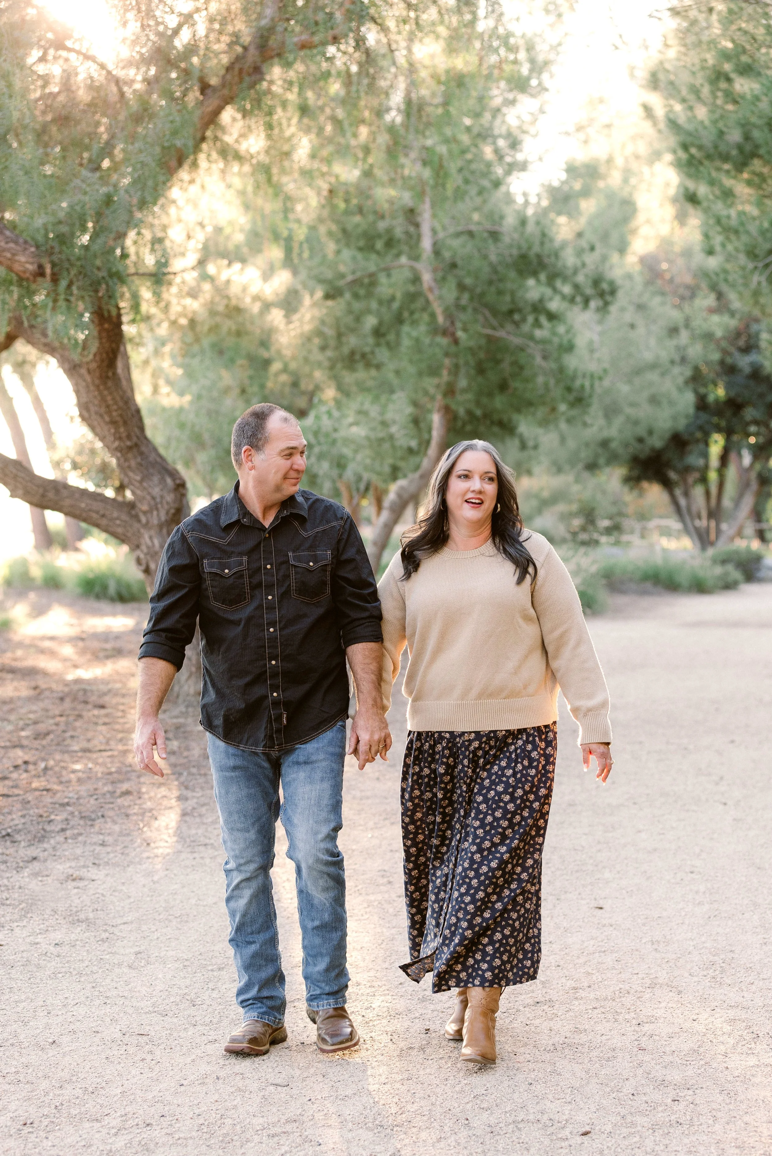 A couple walking hand in hand outdoors on a dirt path surrounded by trees, with sunlight filtering through the leaves.