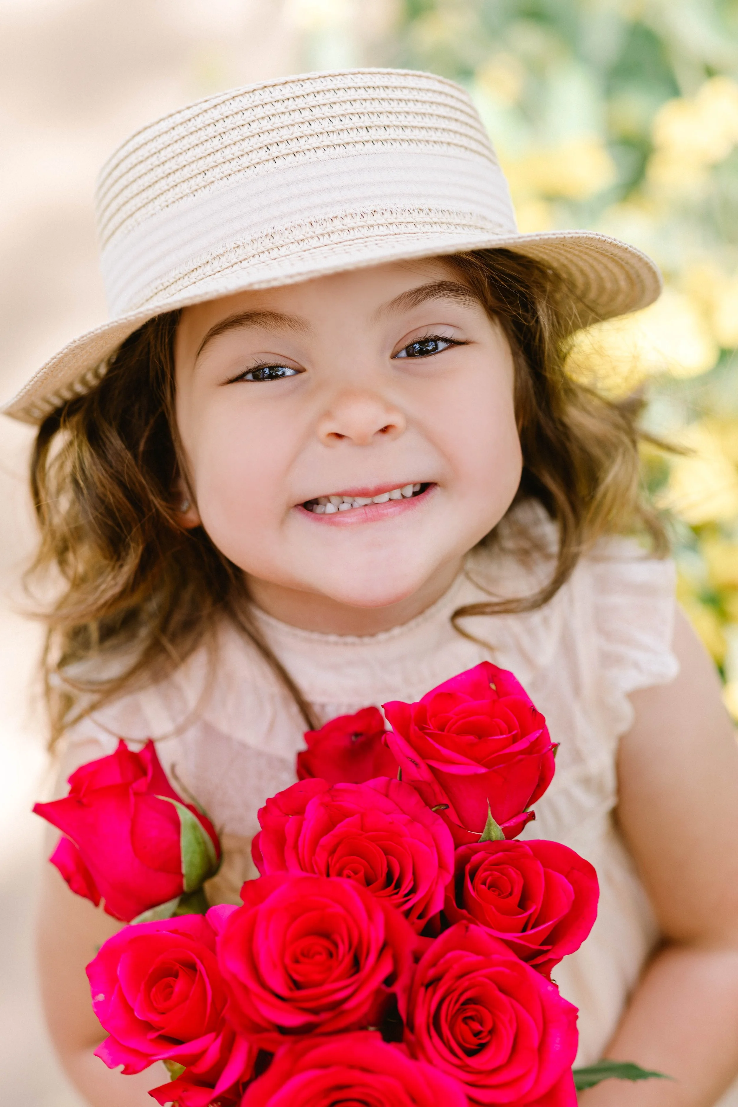 A young girl with brown curly hair wearing a beige hat and a light-colored top, holding a bouquet of red roses, smiling at the camera outdoors.