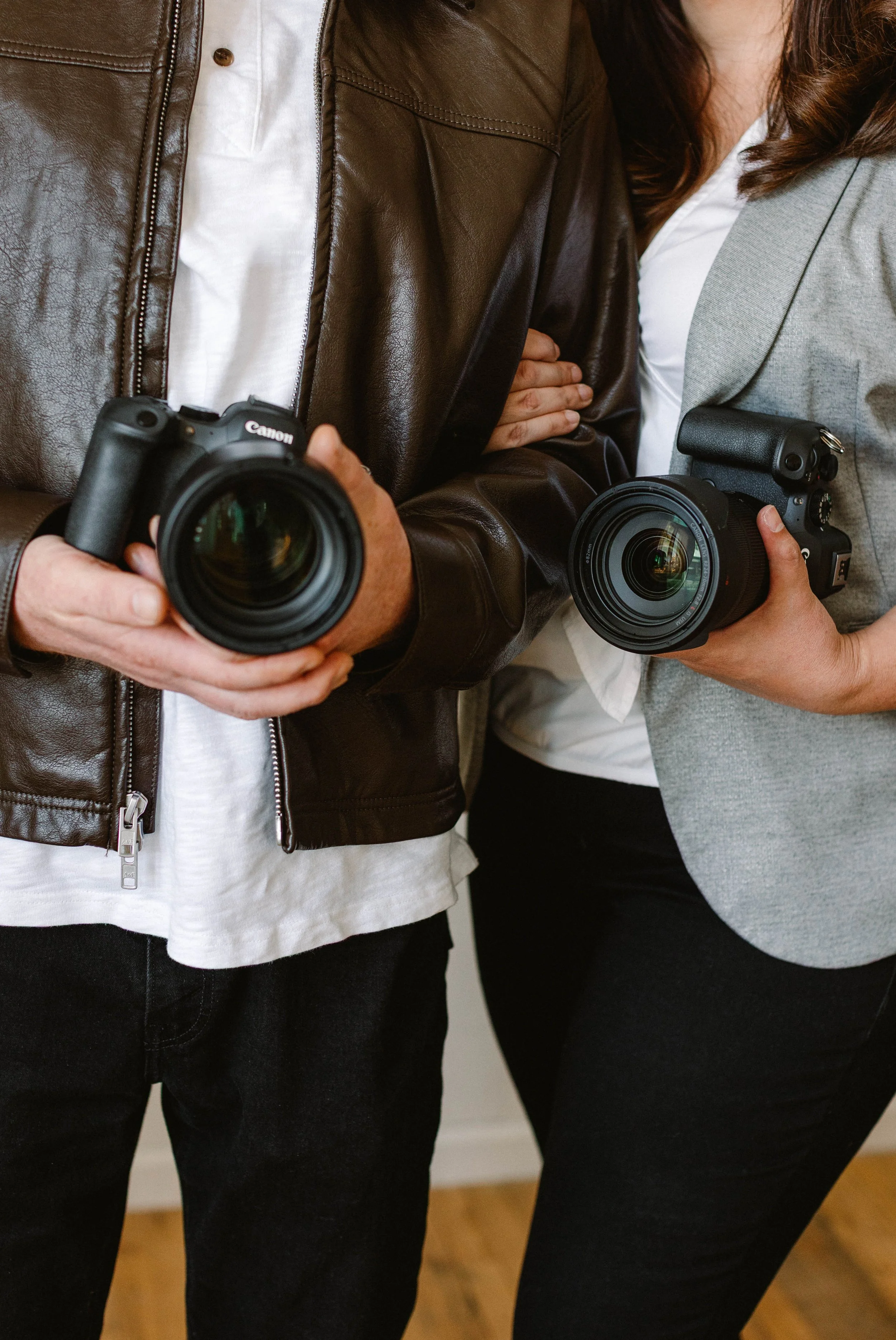 Two people holding professional cameras, standing close together indoors, with one person wearing a brown leather jacket and the other a gray blazer.