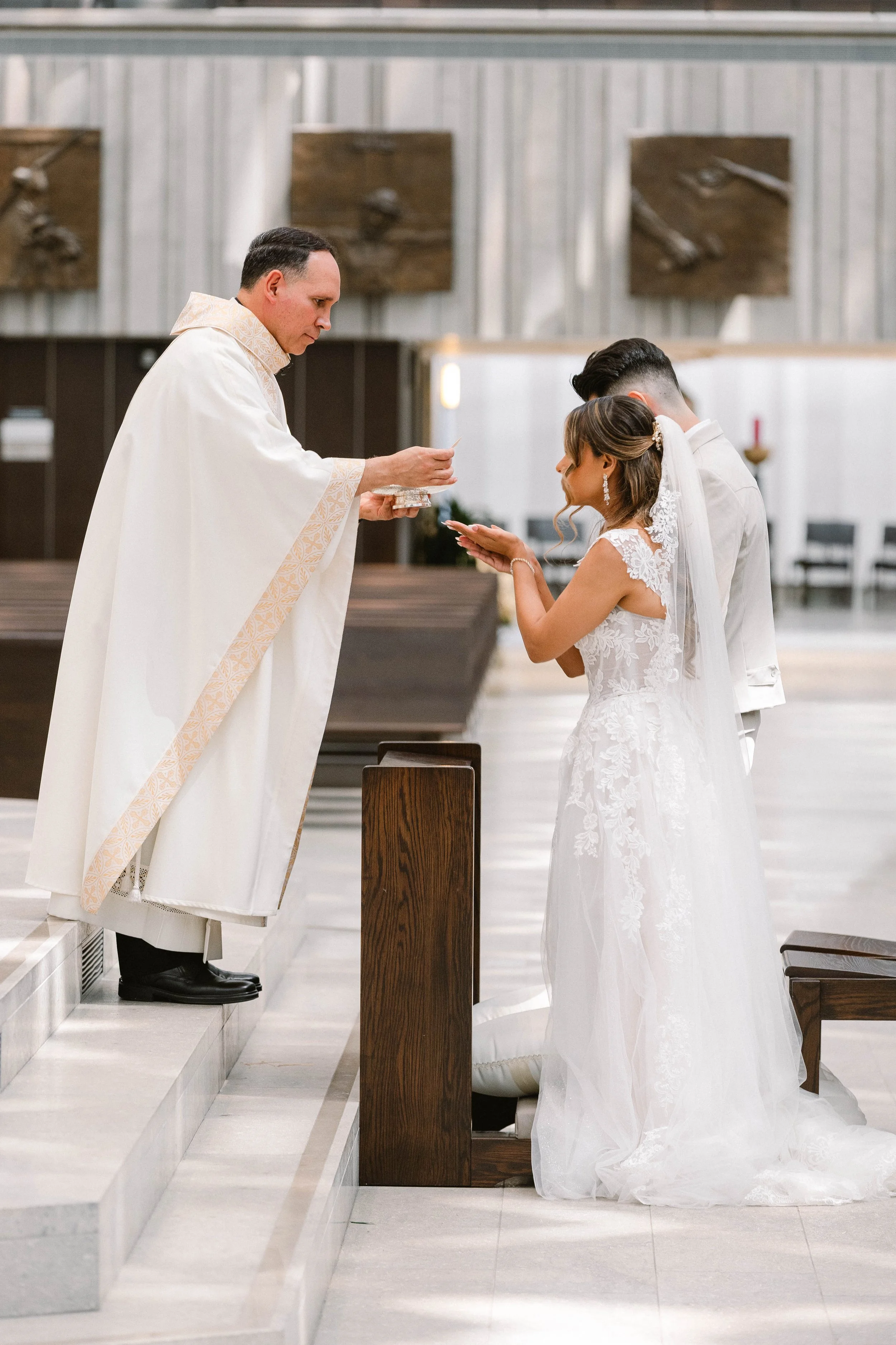 A bride and groom kneeling in front of a priest during a wedding ceremony in a church, with the priest holding a card or paper.
