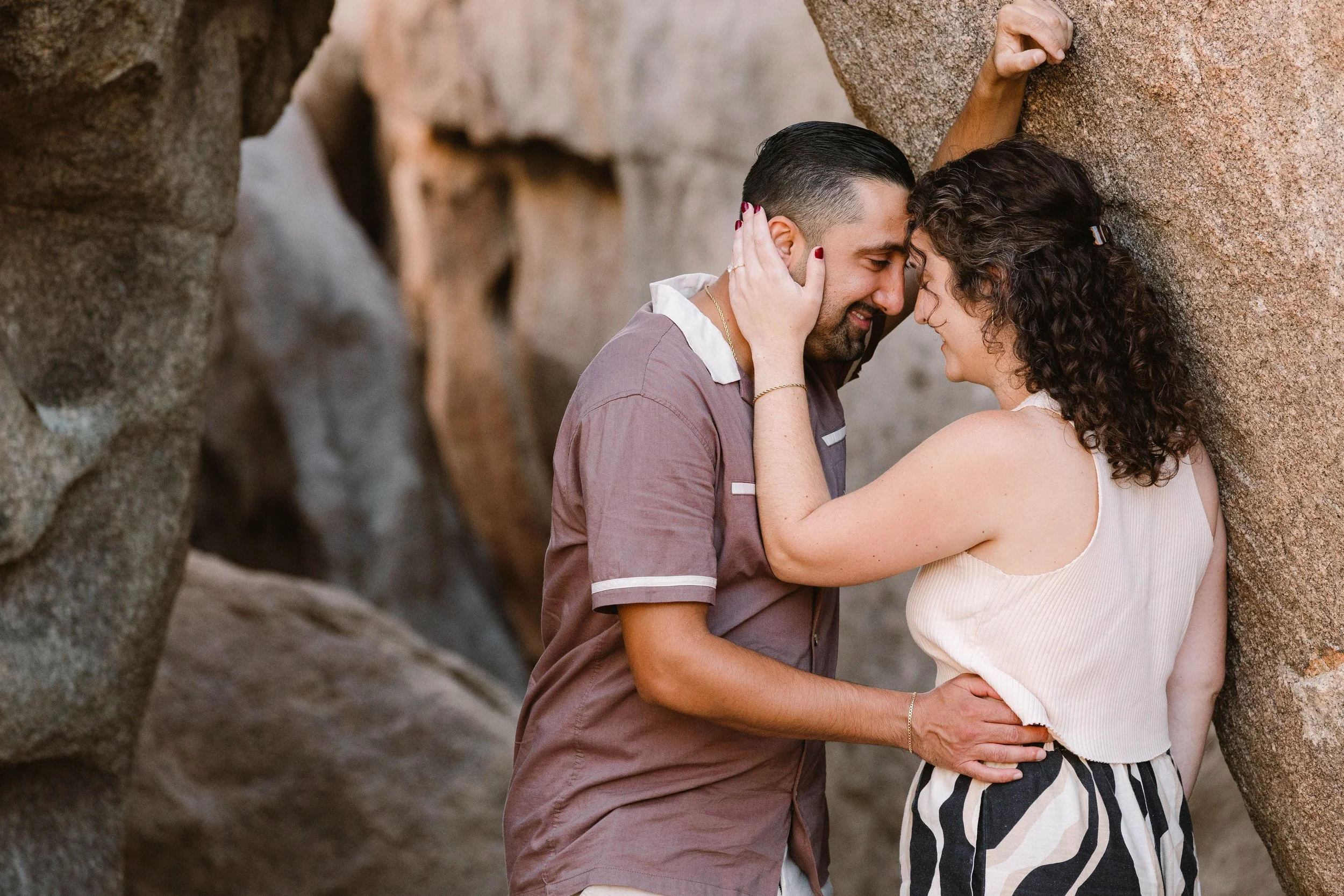 A couple sharing an intimate moment with foreheads touching, standing between large rocks outdoors.