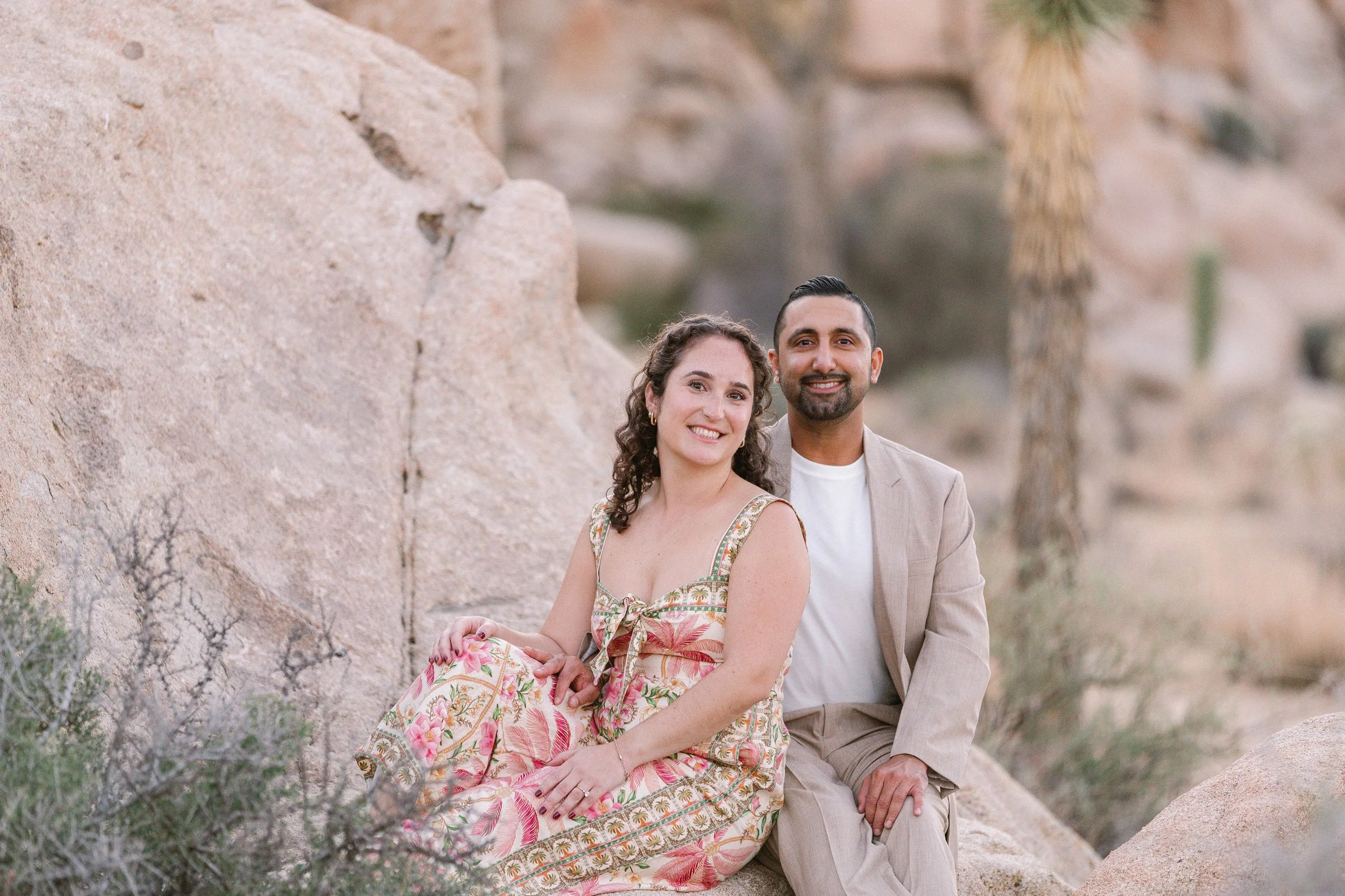 A smiling woman in a floral dress and a man in a beige suit sitting on rocks in a desert landscape with rocks and desert plants.