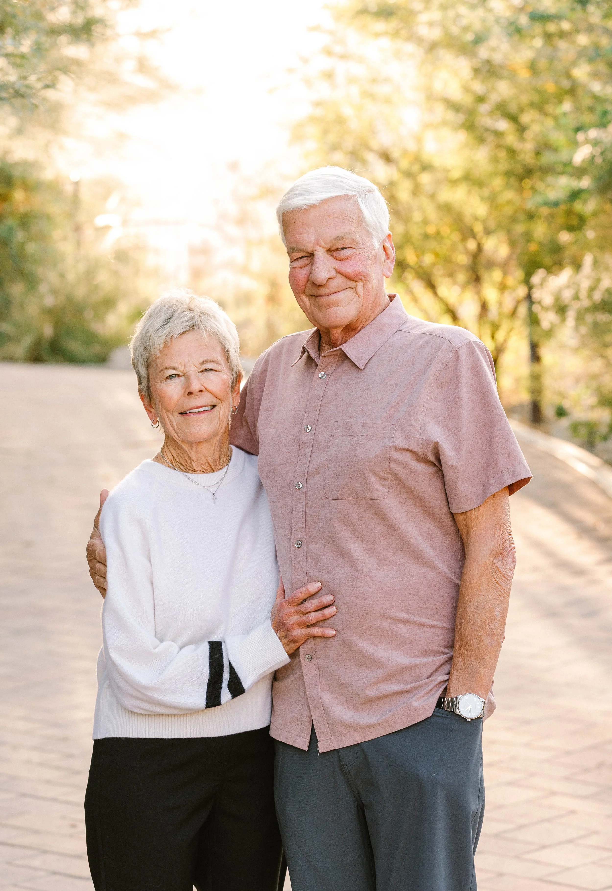 Older couple standing outdoors on a sunny day, smiling and hugging, surrounded by trees with autumn foliage.
