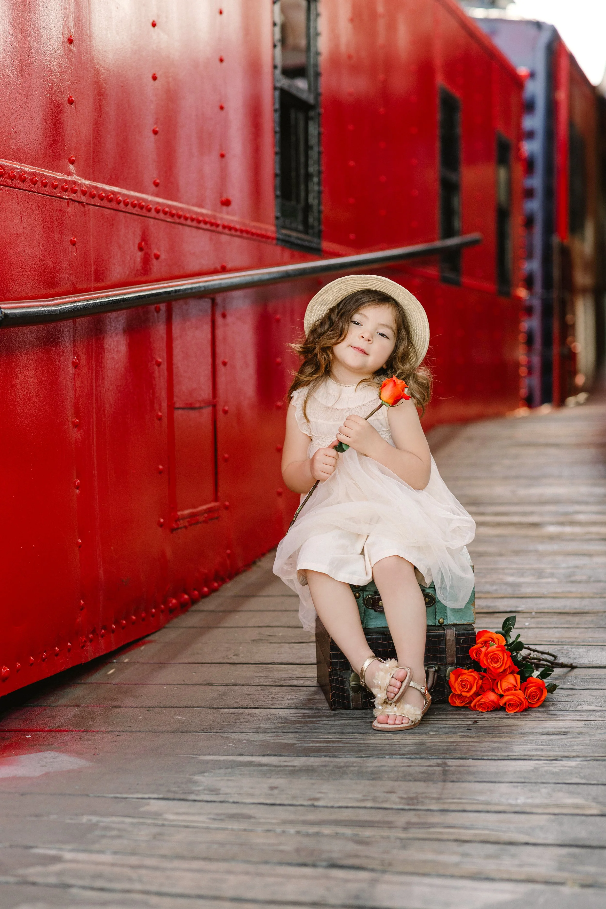 Young girl sitting on a vintage suitcase on a wooden train platform, holding an orange rose, dressed in a white dress and sun hat, with a bouquet of orange roses on the ground beside her.