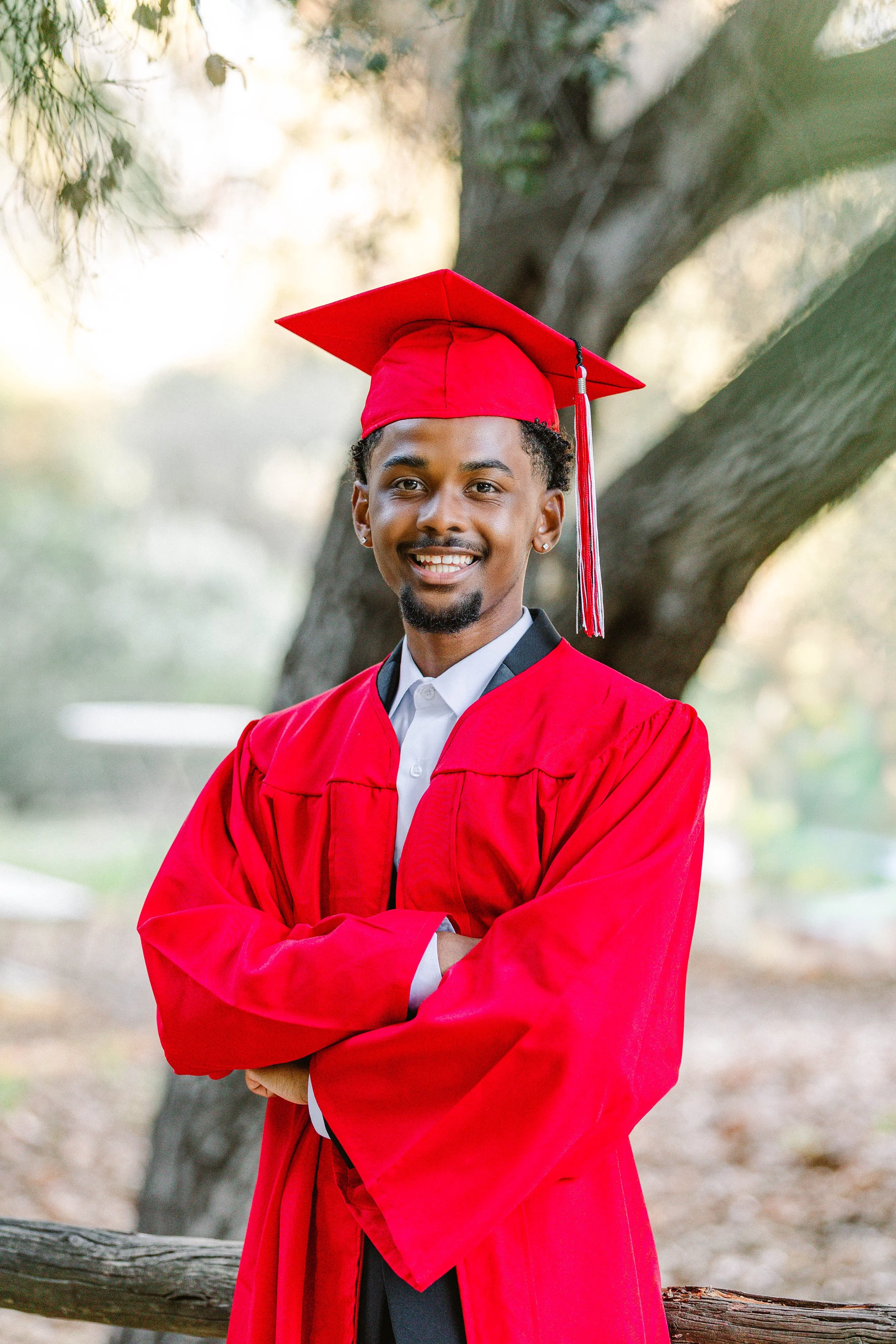 Young man in a red graduation gown and cap standing outdoors with arms crossed and smiling in front of a large tree.