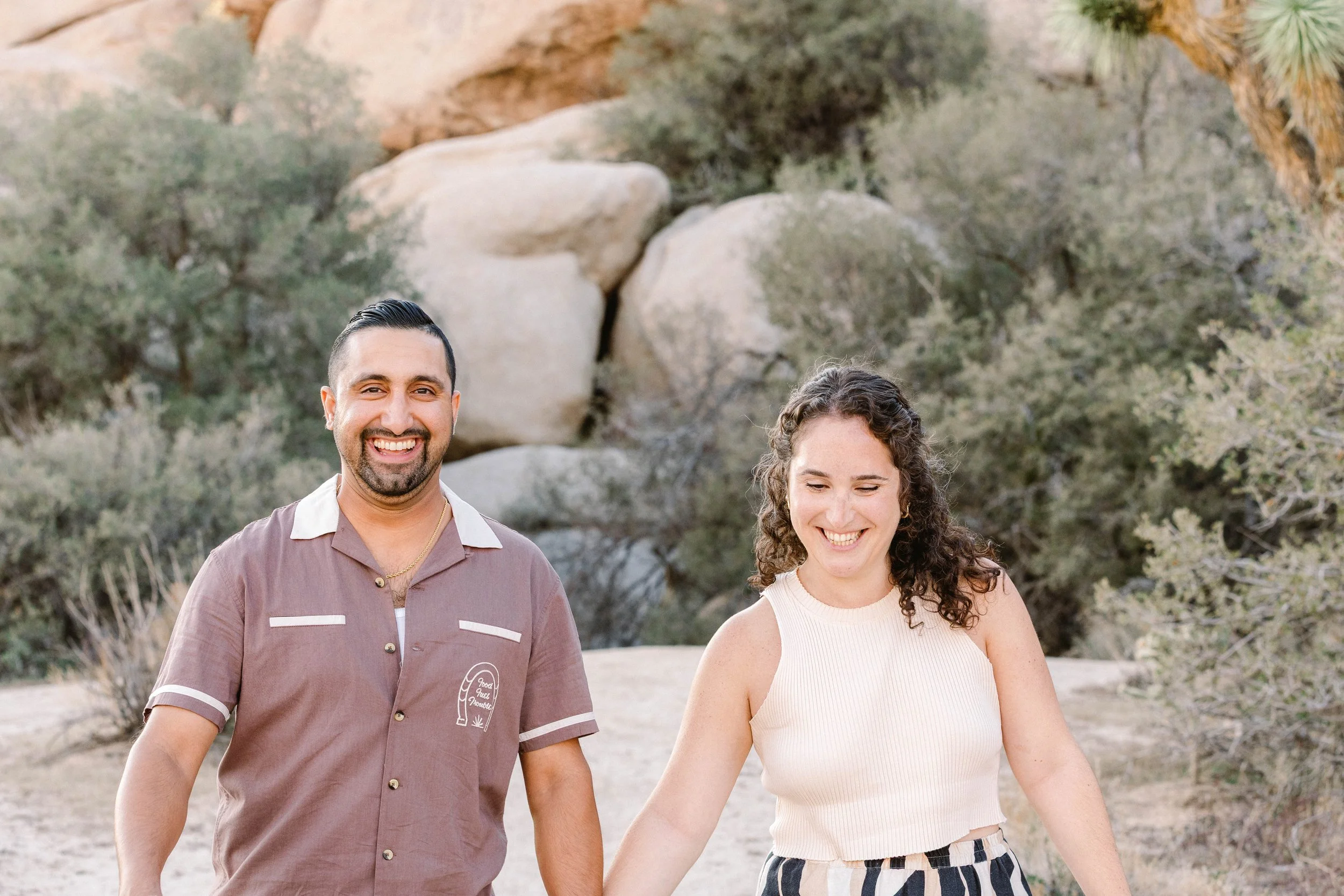 A smiling man and woman walking hand in hand outdoors in a desert landscape with rocks and bushes.