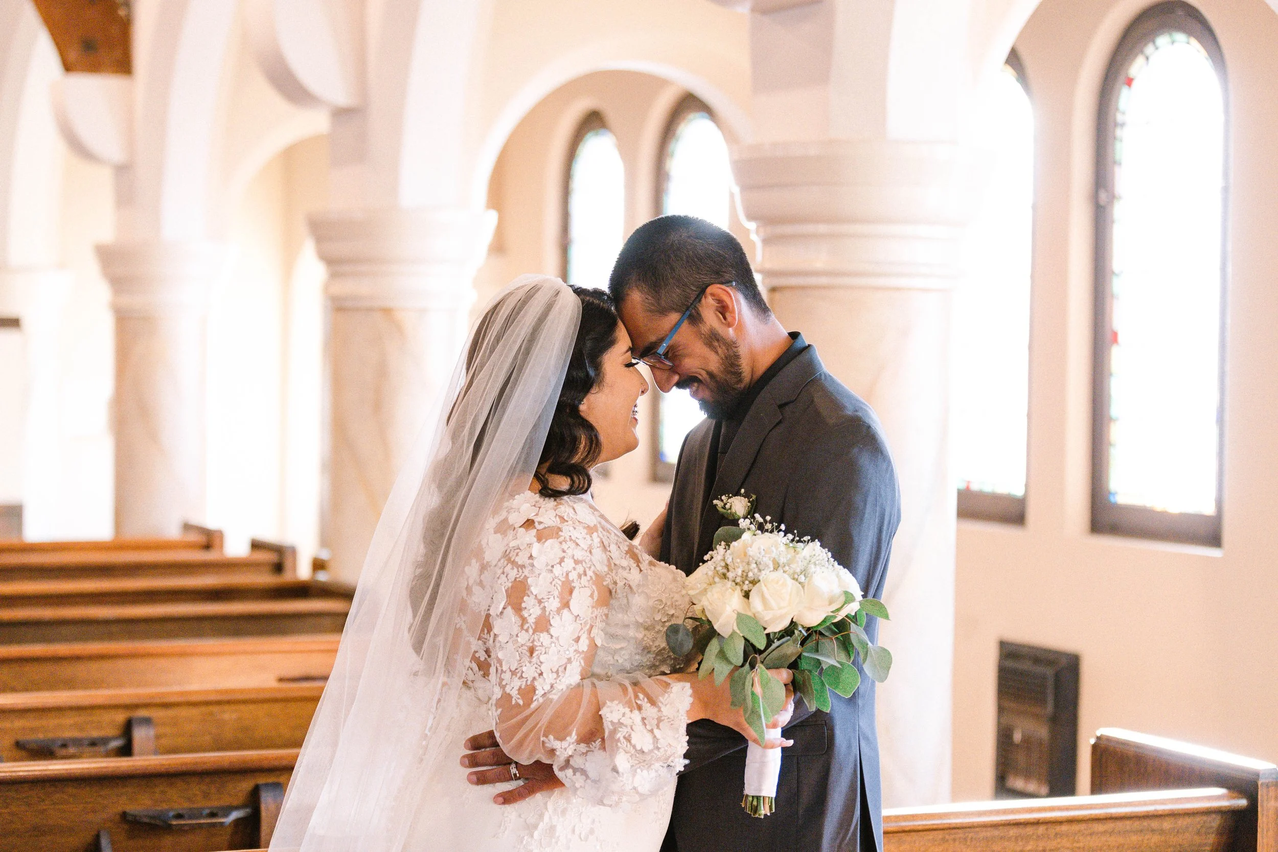 A bride and groom standing close together in a church with stained glass windows, smiling and touching foreheads, the bride holding a bouquet of white roses and greenery.