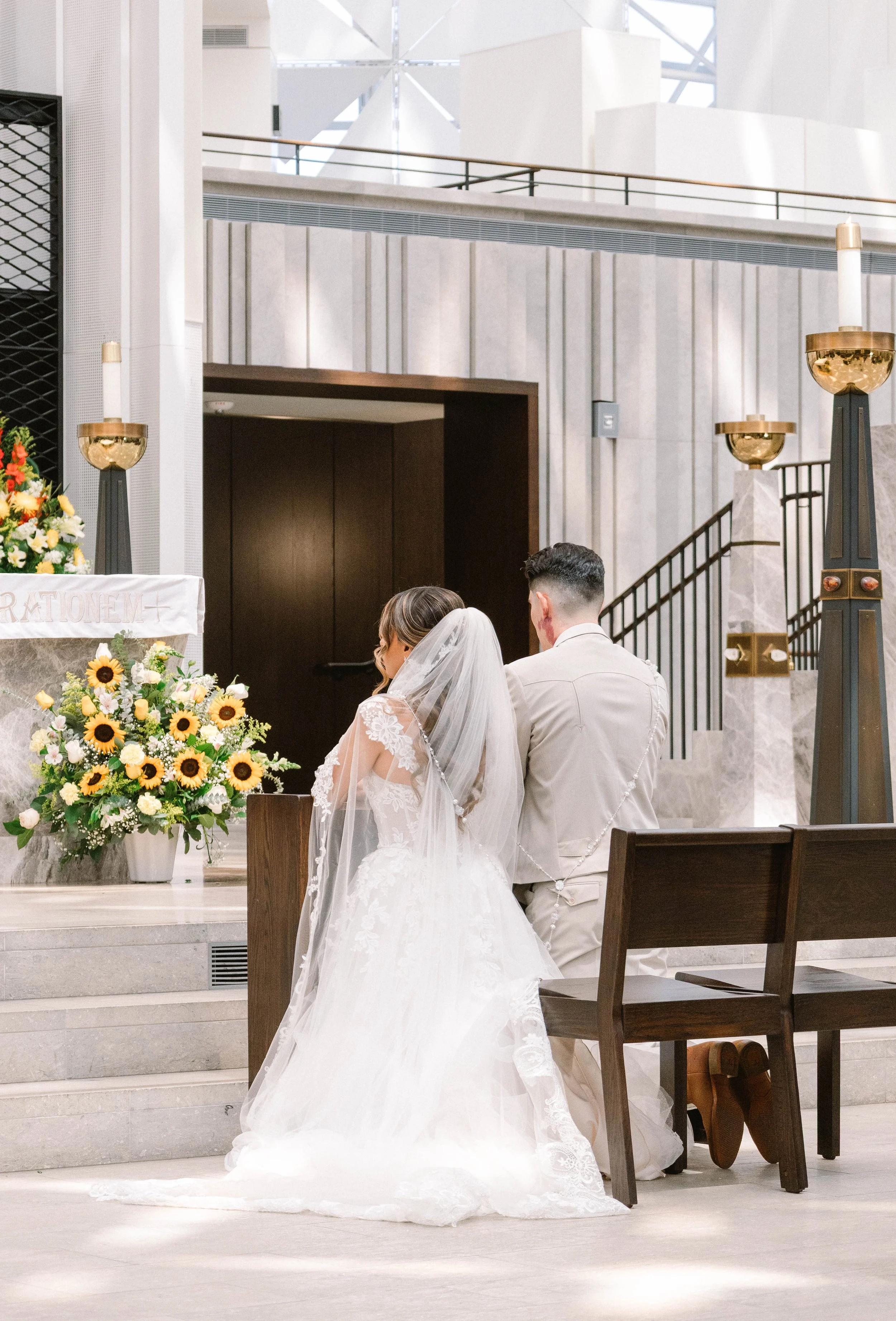 A bride and groom kneeling at the altar during their wedding ceremony inside a modern church with floral arrangements and religious symbols.