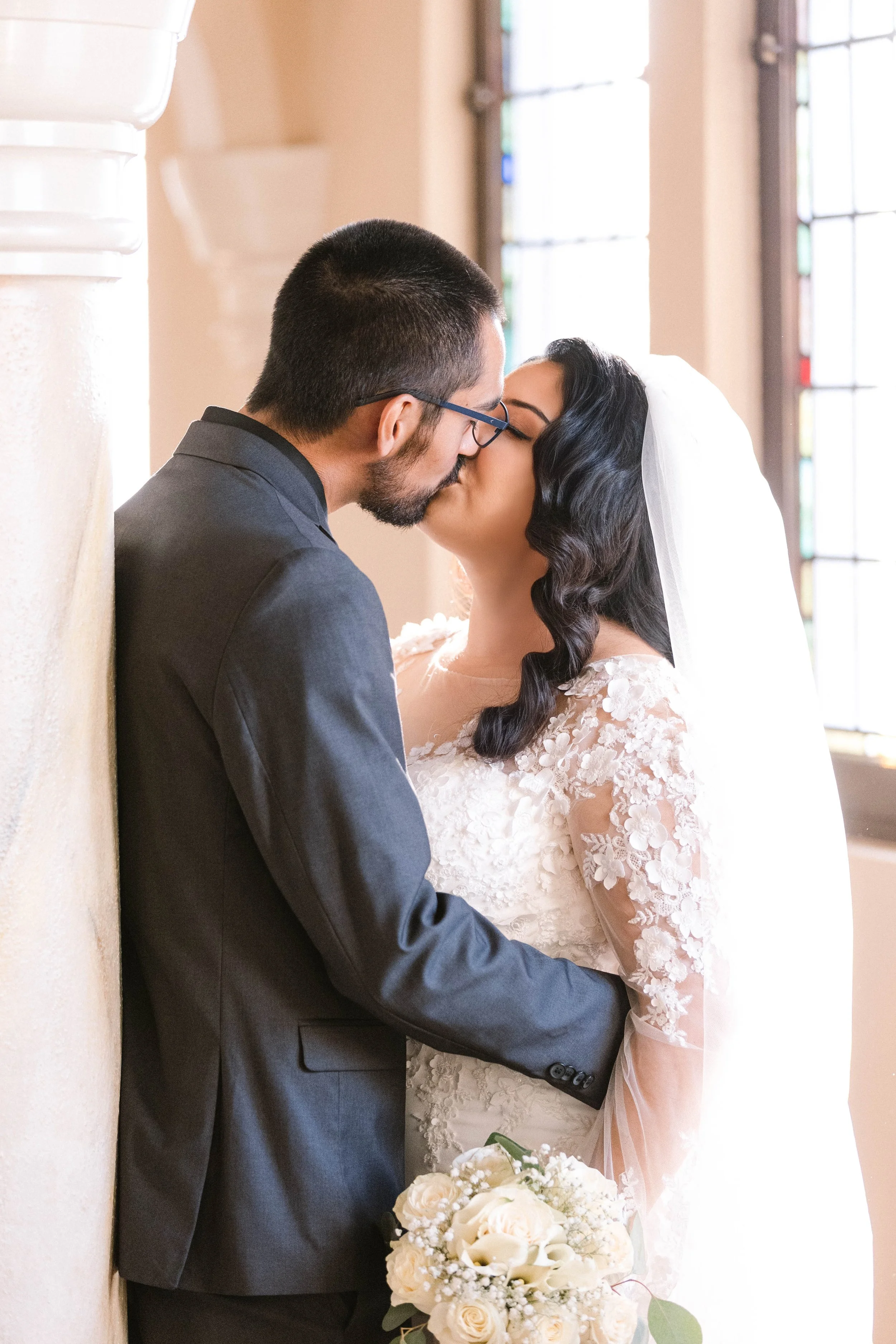 A bride and groom share a kiss indoors near large stained glass windows, with the bride holding a bouquet of white roses and wearing a lace wedding dress, while the groom wears a dark suit.