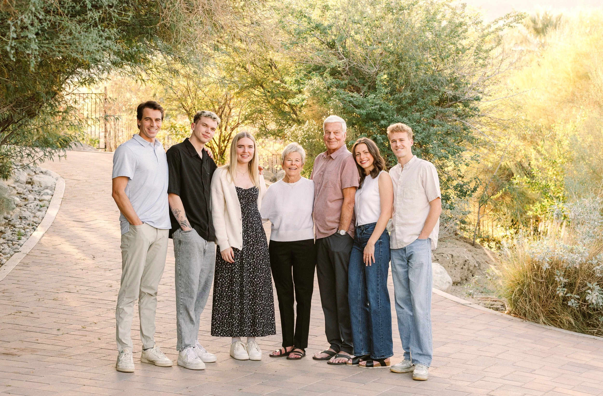 A multigenerational family standing together outdoors on a brick pathway, surrounded by trees and sunlight.